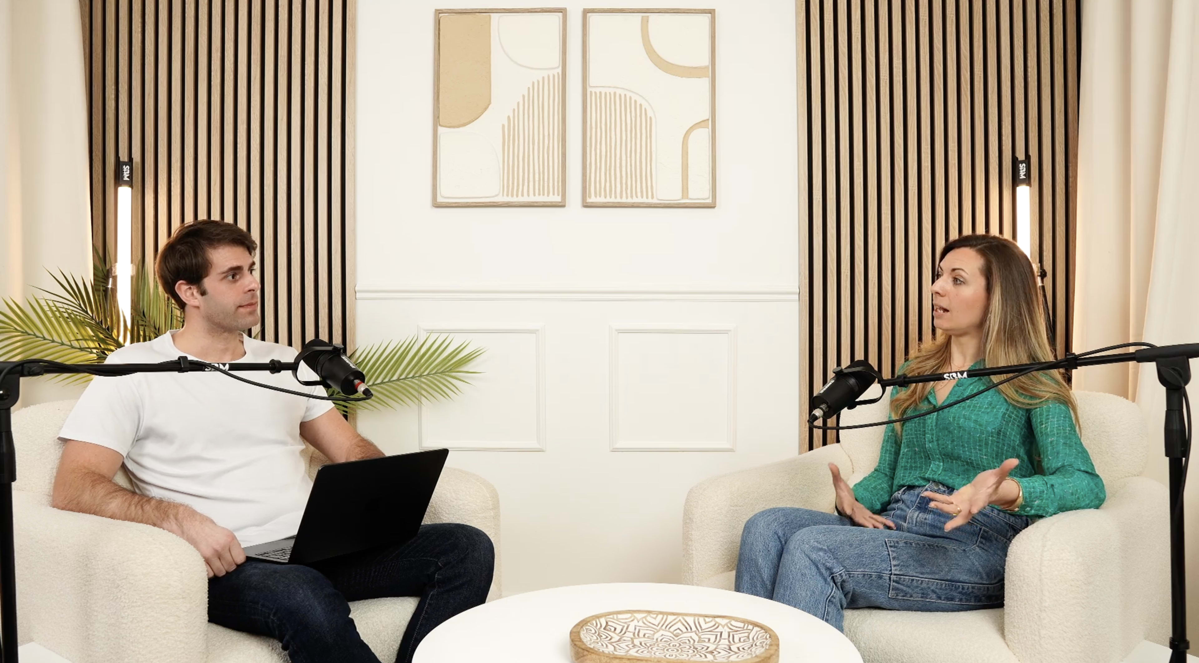 A man and a woman are seated in a modern, minimalist room, engaged in conversation with microphones placed on stands in front of them.