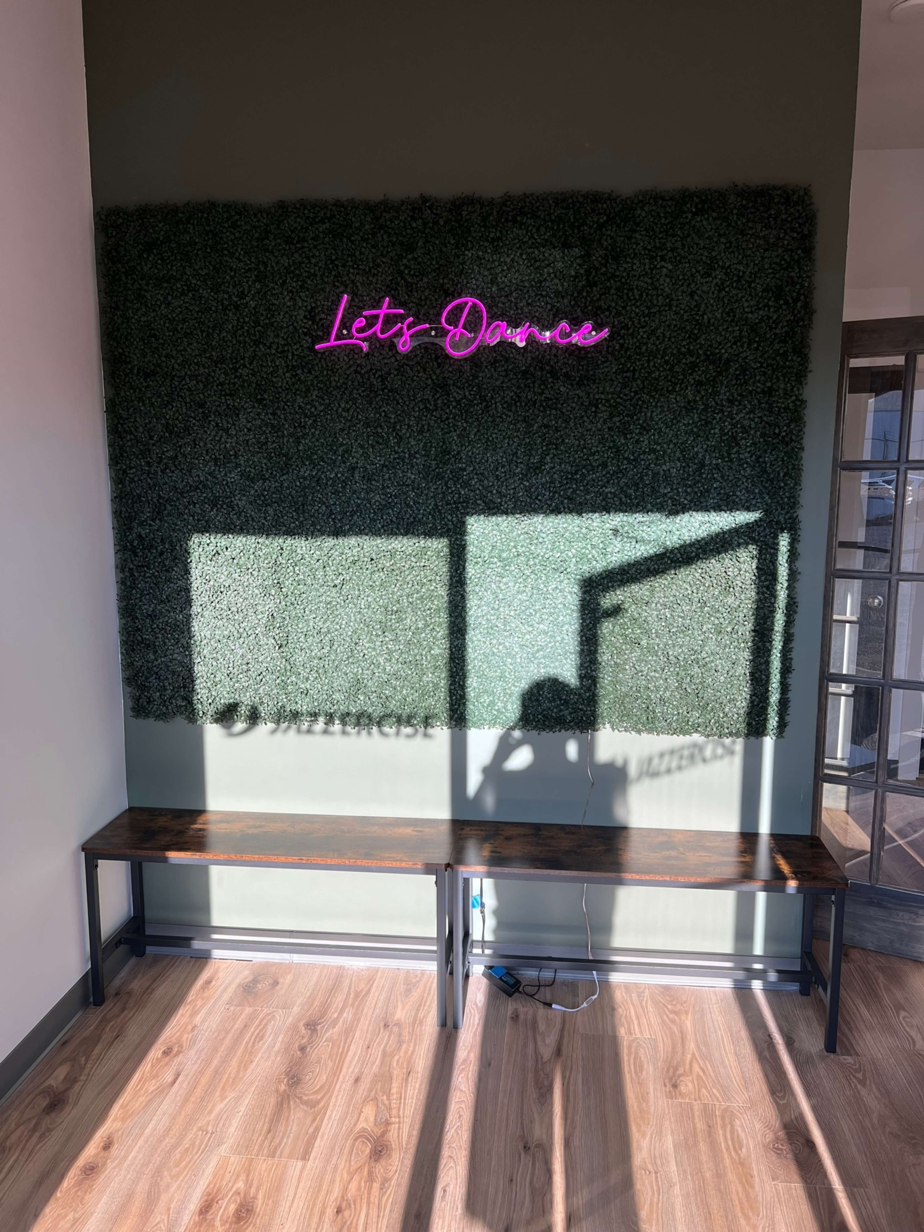 A neon sign reading "Let's Dance" is mounted on a textured green wall, with shadows cast onto a wooden bench in the foreground.