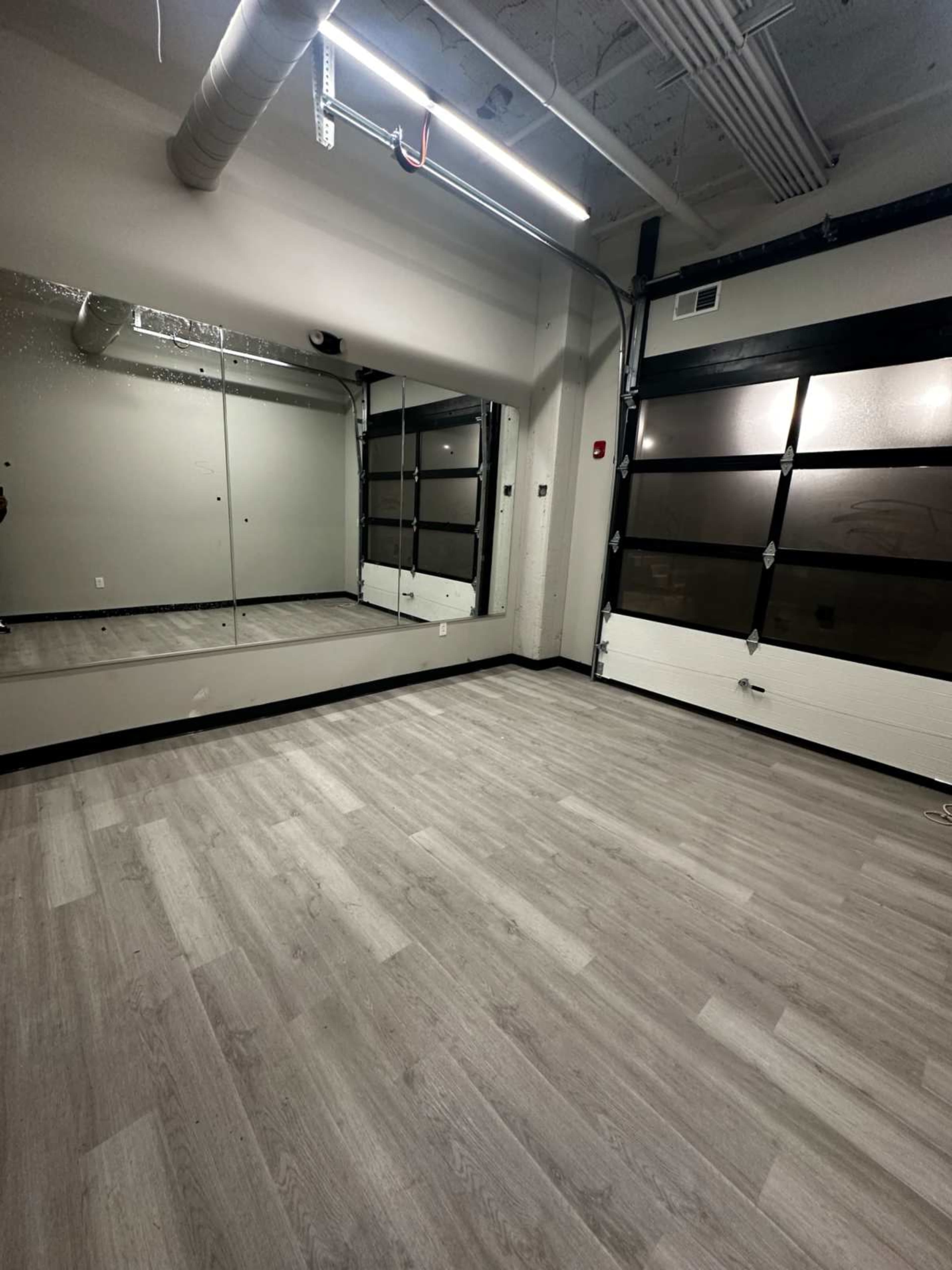 The image shows an empty room with grey wooden flooring, a large mirror on the wall, and a garage-style rolling door.
