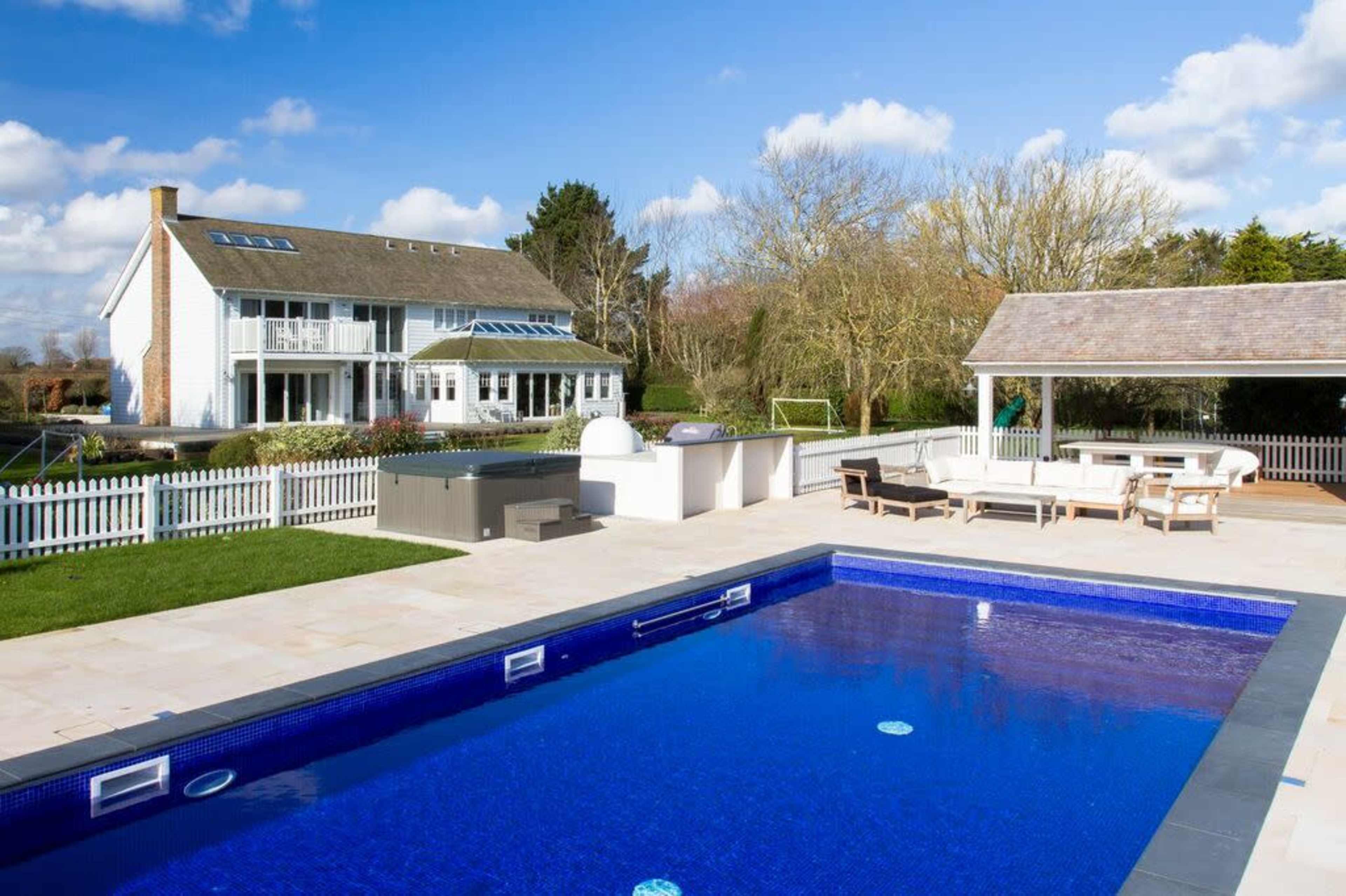 A backyard with a blue swimming pool, a large house in the background, and a patio area with seating and an outdoor kitchen.