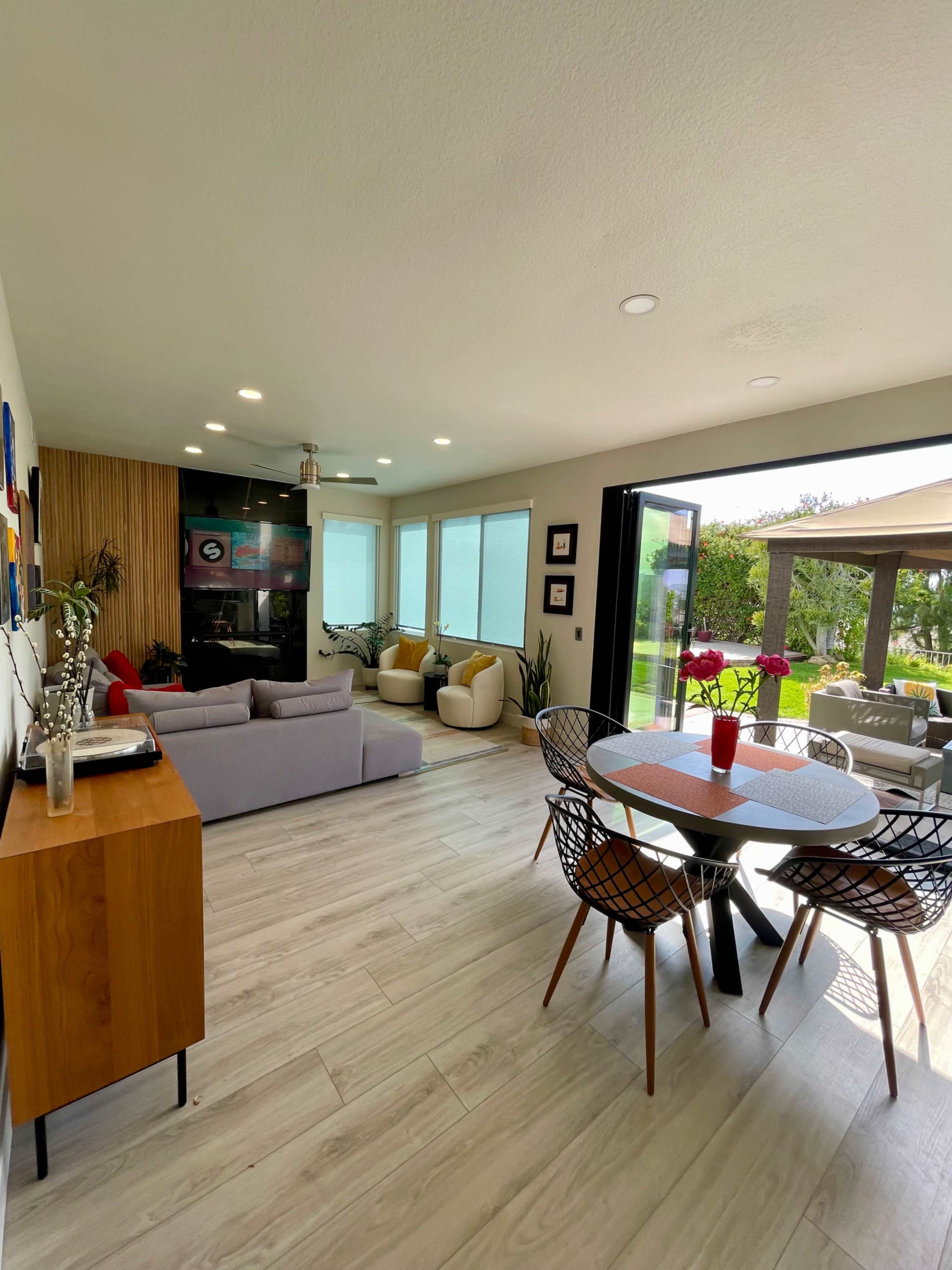 A bright, modern living room with a sofa facing a television, a dining area with a round table, and large sliding glass doors opening to an outdoor patio.