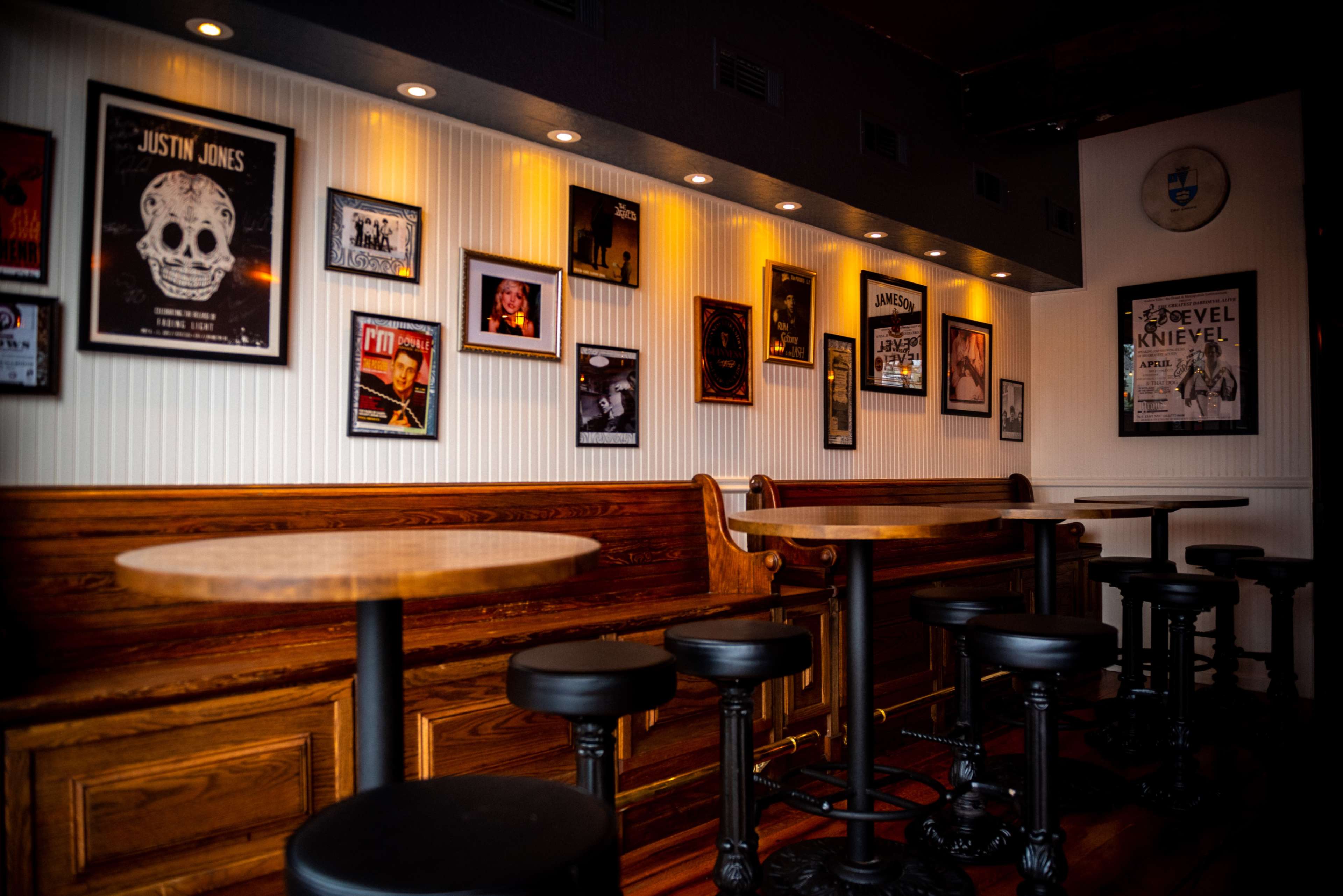 The image shows a dimly lit bar interior with wooden benches and tall stools, featuring framed music and event posters on the walls.