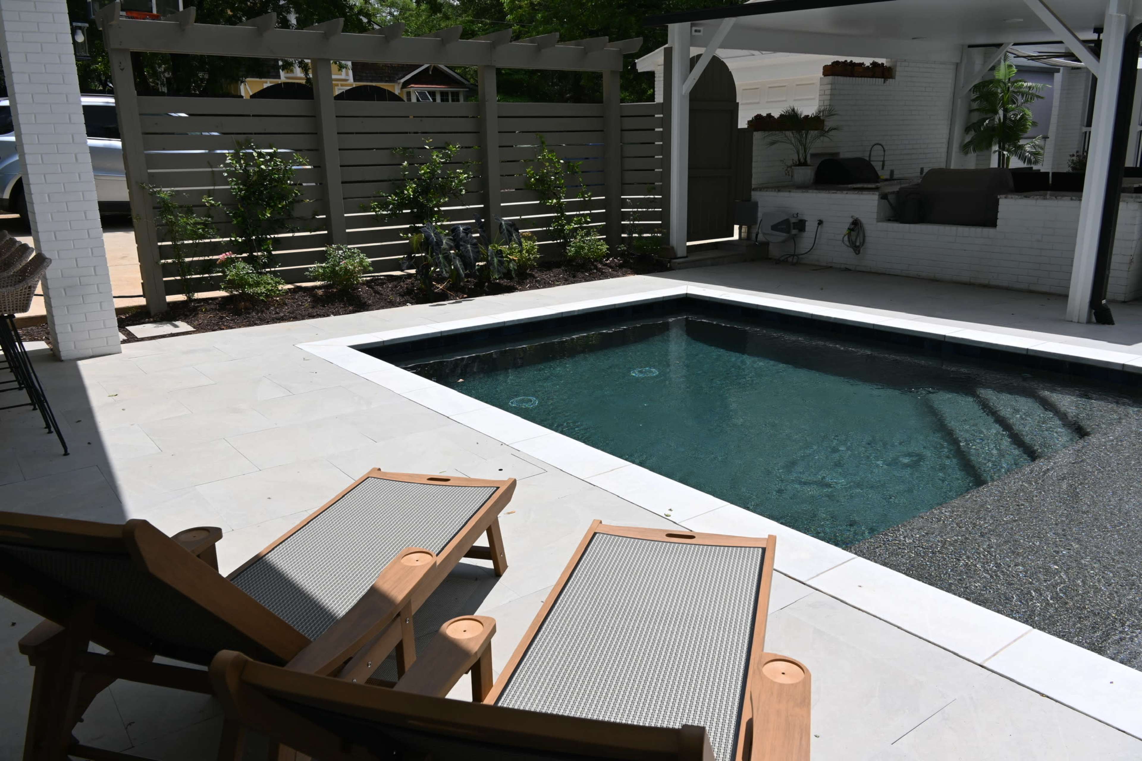 The image shows a modern outdoor space featuring a swimming pool surrounded by patio tiles and two wooden lounge chairs.
