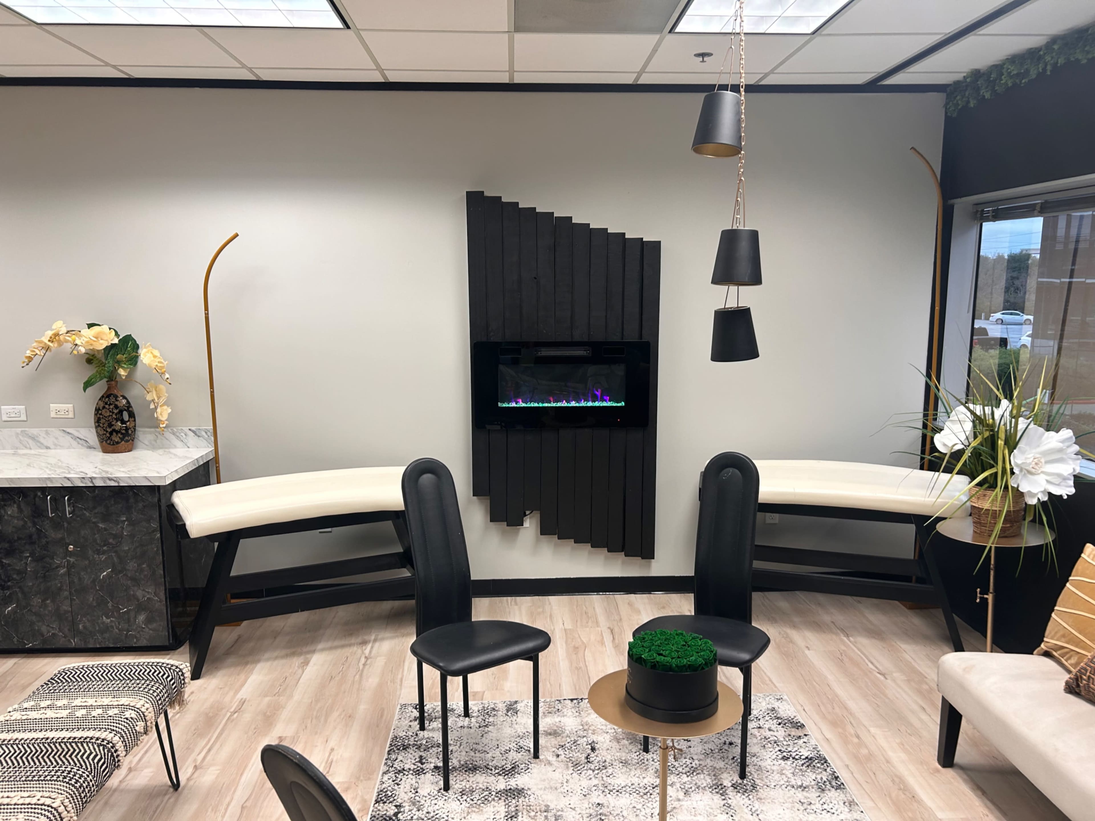 The image shows a modern waiting area featuring a wall-mounted electric fireplace, two black chairs, a small round table, and decorative elements like plants and flowers.