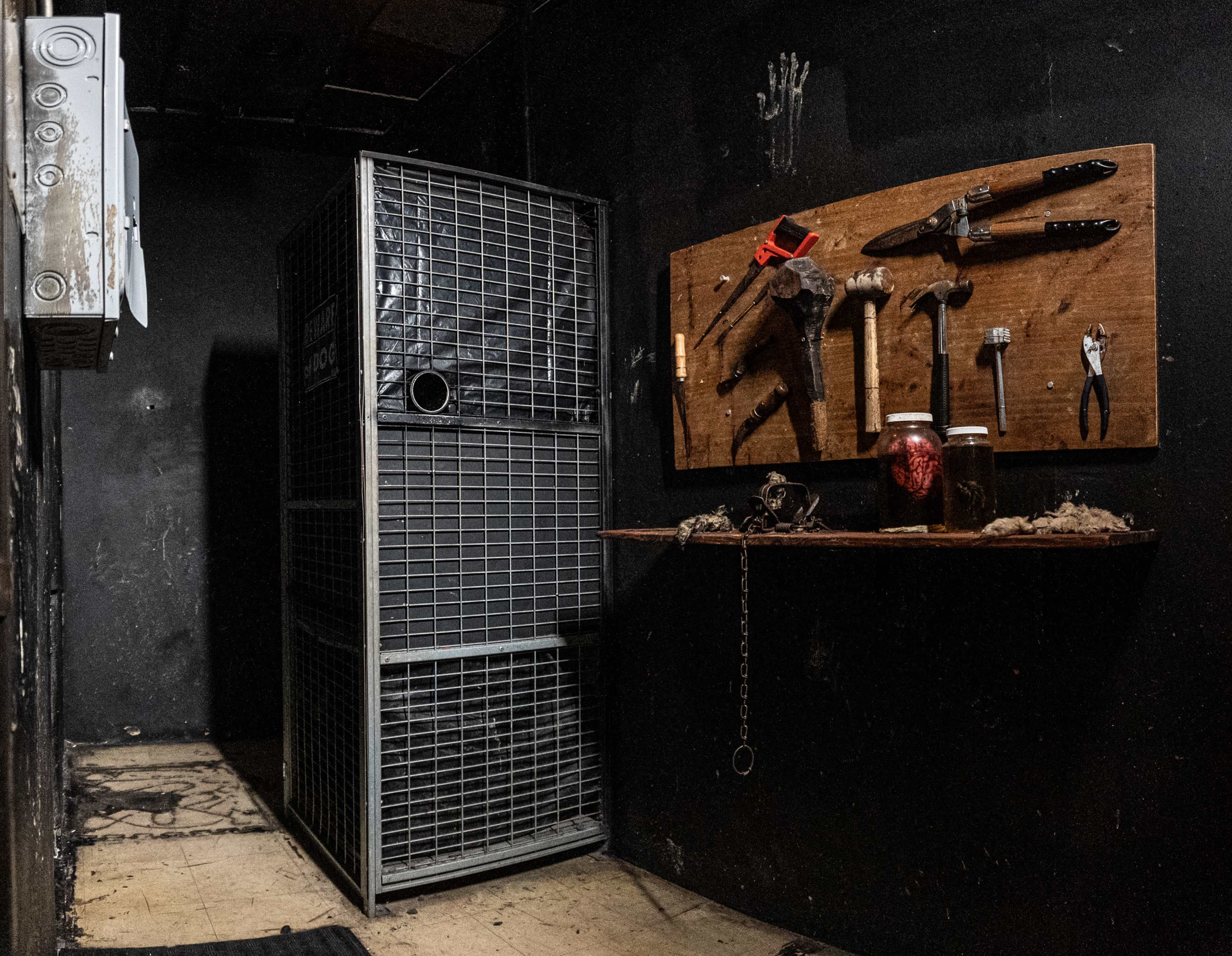 A dimly lit room with a large metal cage on the left and a wall-mounted board displaying various tools on the right.