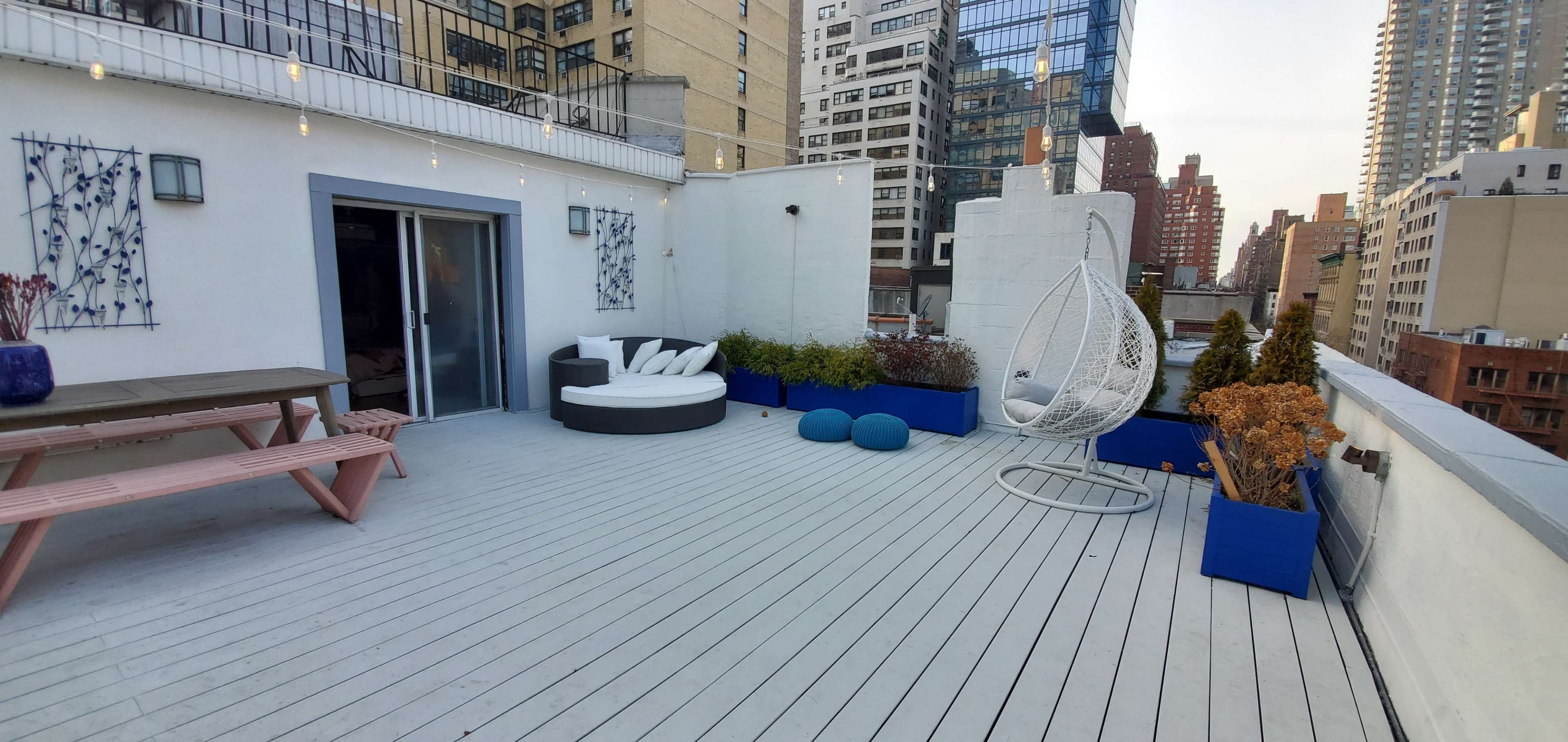 A spacious rooftop terrace featuring seating areas, colorful planters, and city buildings in the background.