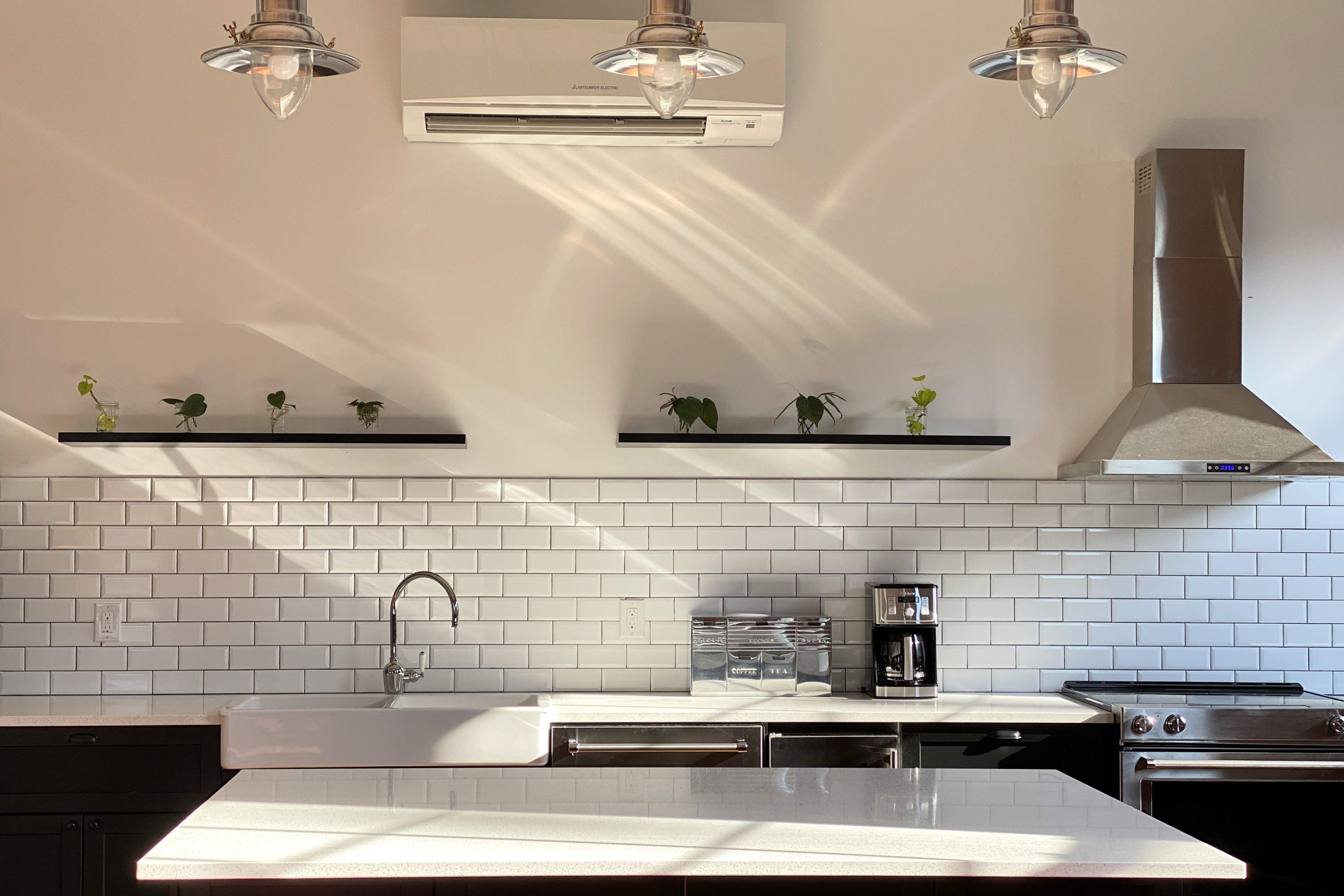 A modern kitchen with white tiles, a large sink, a stovetop, and shelves displaying small plants under overhead lighting.