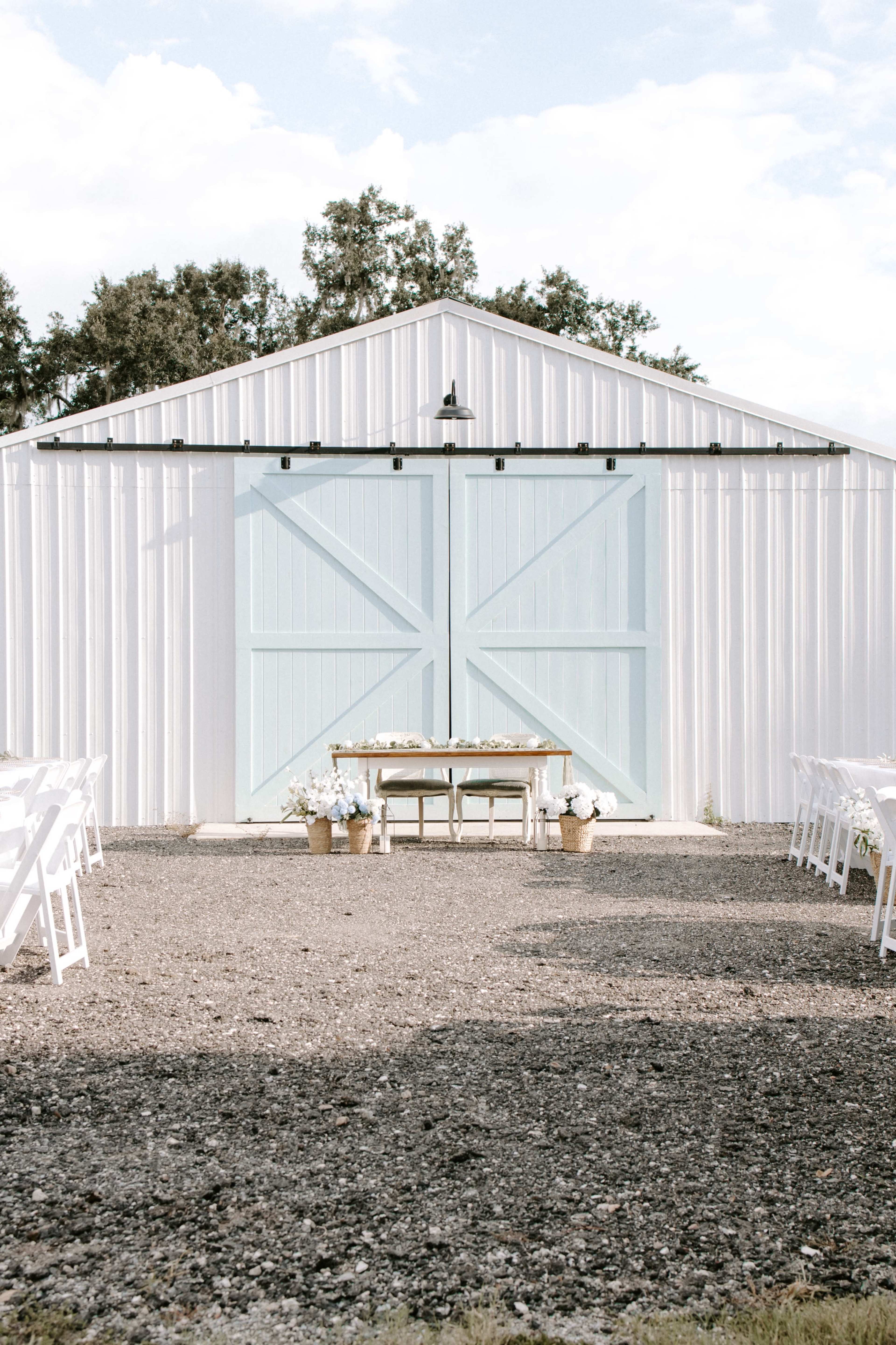 The image shows a serene outdoor wedding setup with a light blue barn door as the backdrop, flanked by rows of white chairs and decorated with potted flowers.