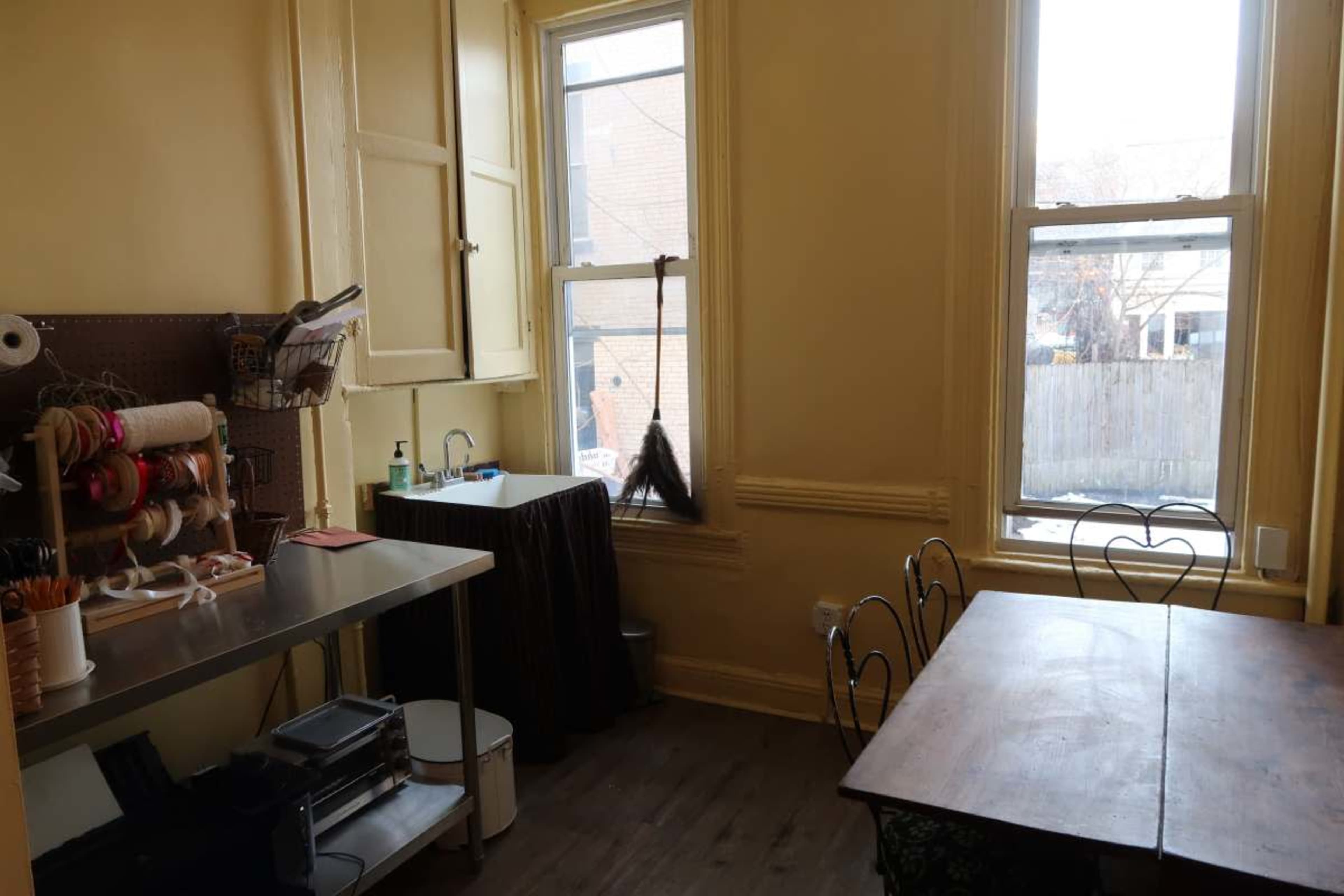 The image shows a small, sunlit room with a metal table, a sink, and a wooden dining table surrounded by chairs.