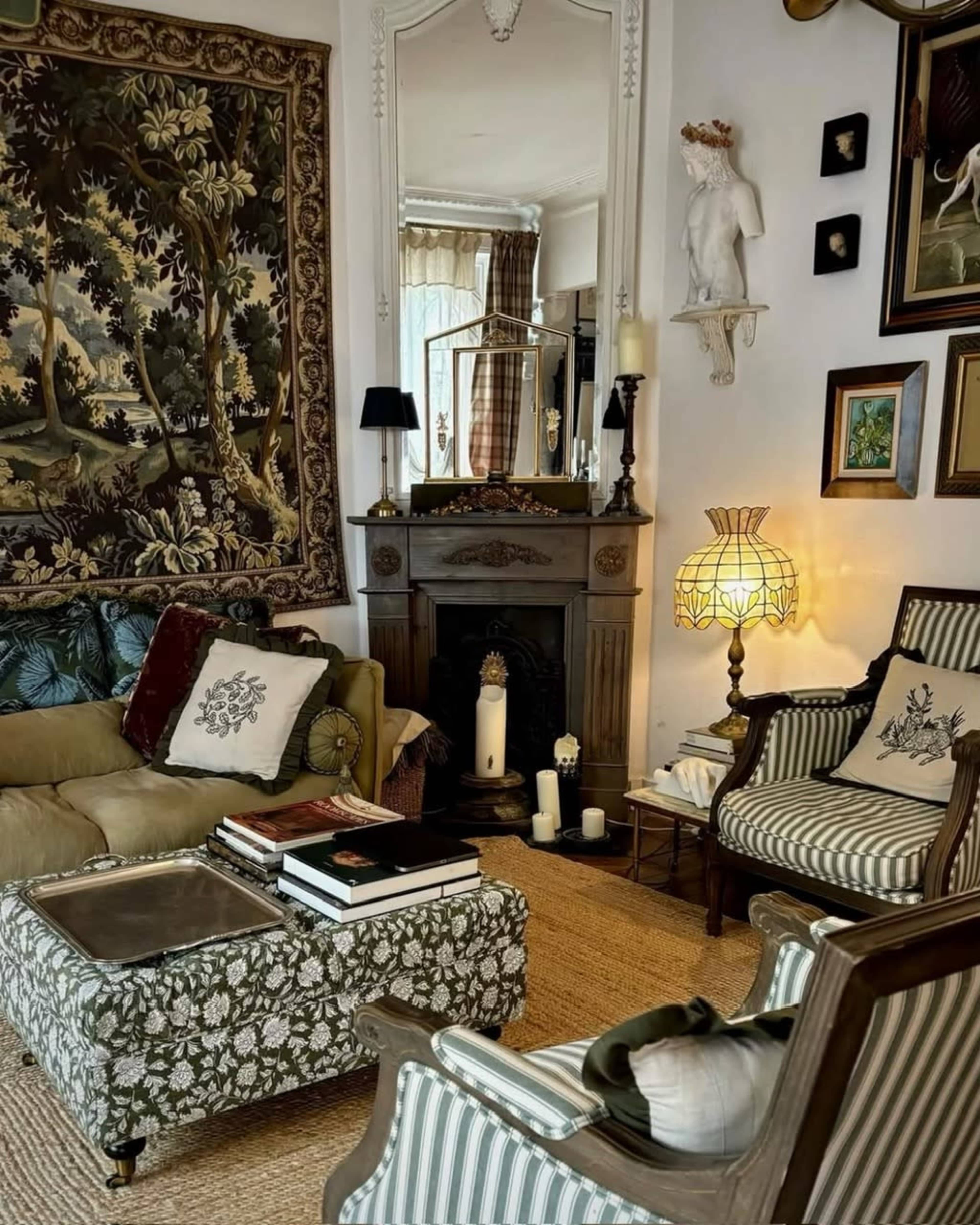 The image shows a cozy living room with a vintage aesthetic, featuring a mix of upholstered furniture, a fireplace with decorative elements, and a large tapestry hanging on the wall.