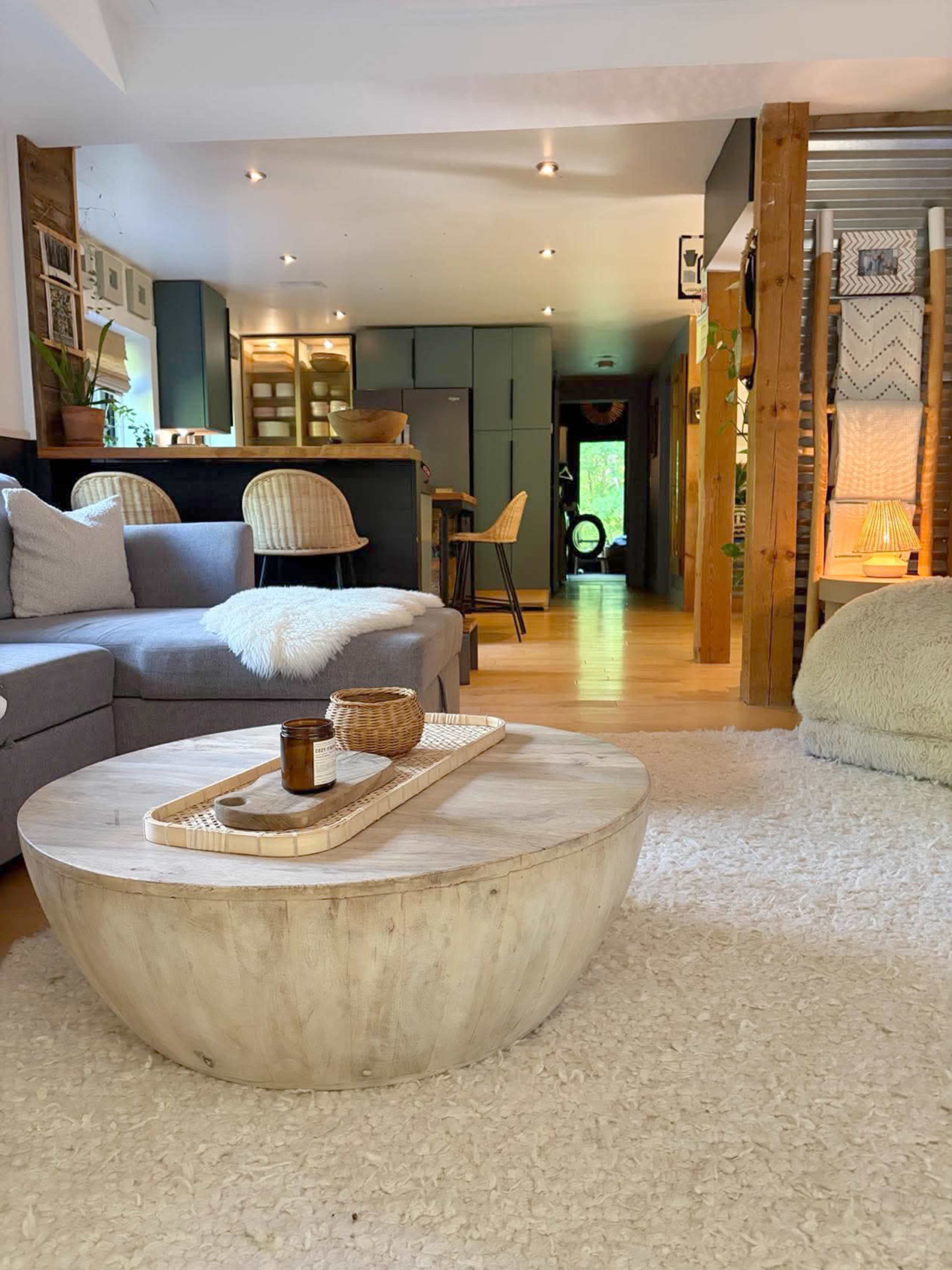 The image shows a cozy living room with a gray sectional sofa, a round coffee table, and a light-colored rug, leading to a kitchen and dining area in the background.