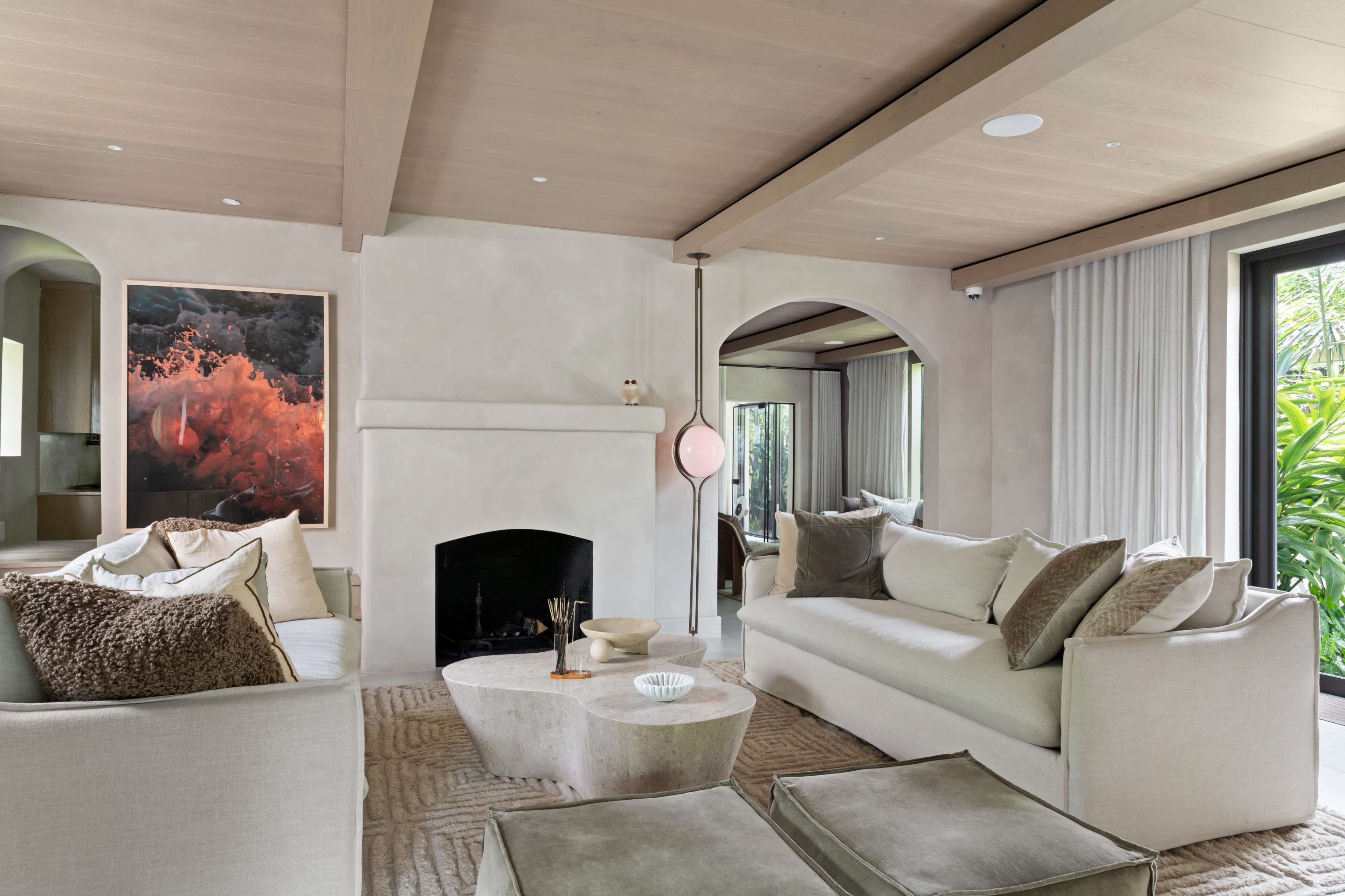 The image depicts a modern living room with a pale color palette, featuring a cozy seating area, a minimalist fireplace, and large windows that offer views of greenery outside.