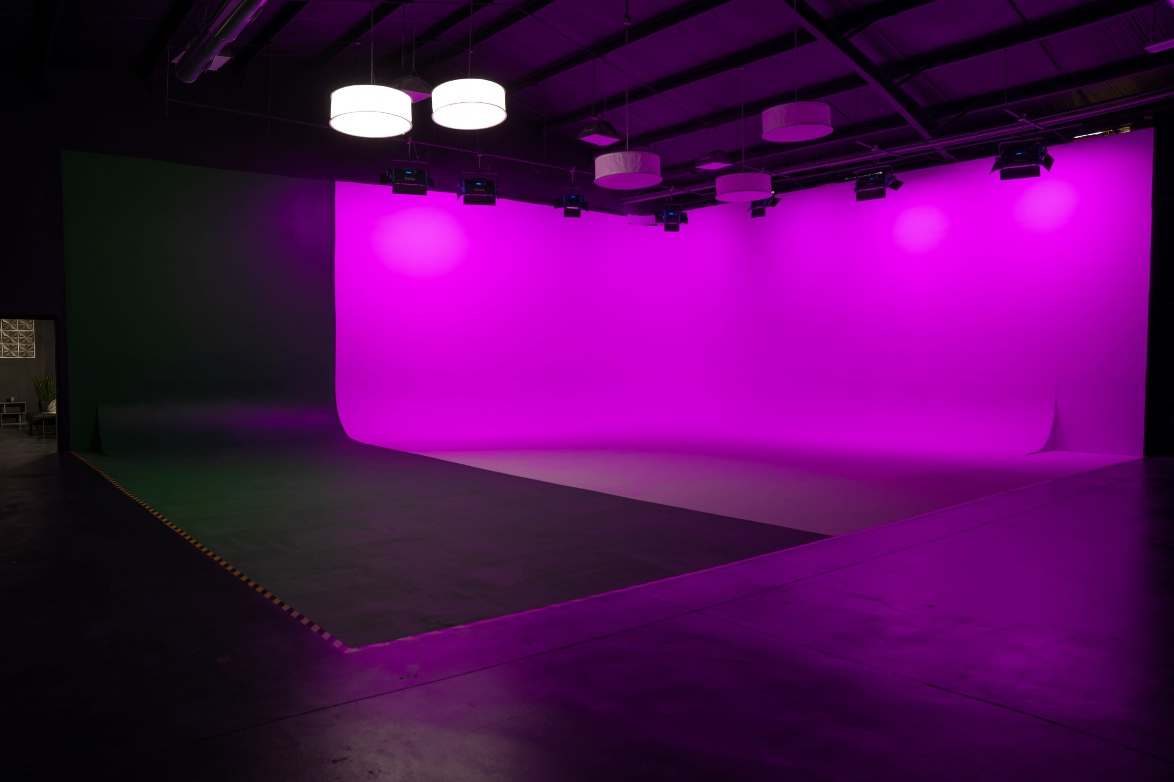 A studio space featuring a large, curved backdrop illuminated in pink, with hanging lights above and a dark floor.