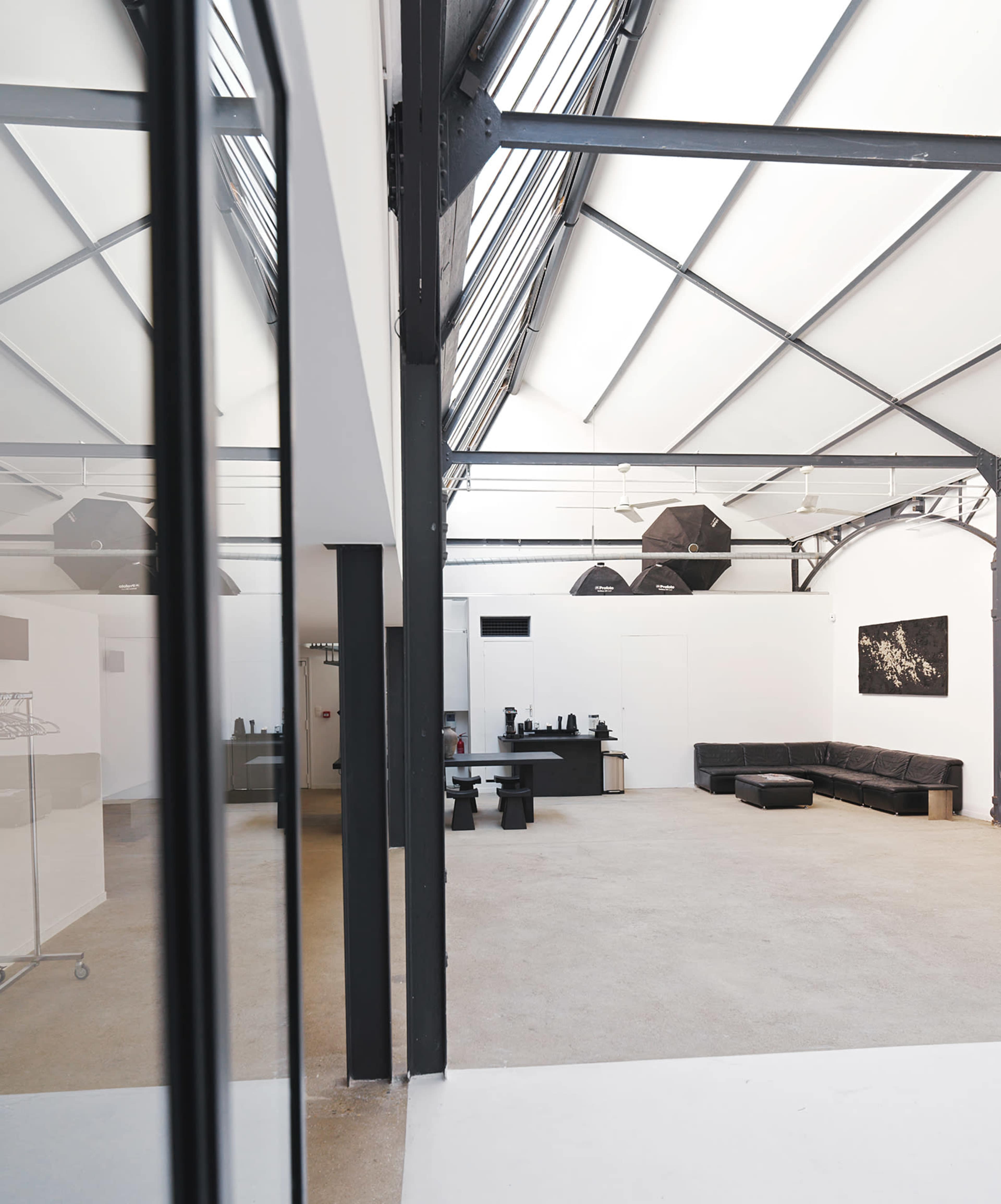 The image shows a spacious, modern studio with high ceilings, large windows, and minimal furnishings, including a black sofa and a work table.