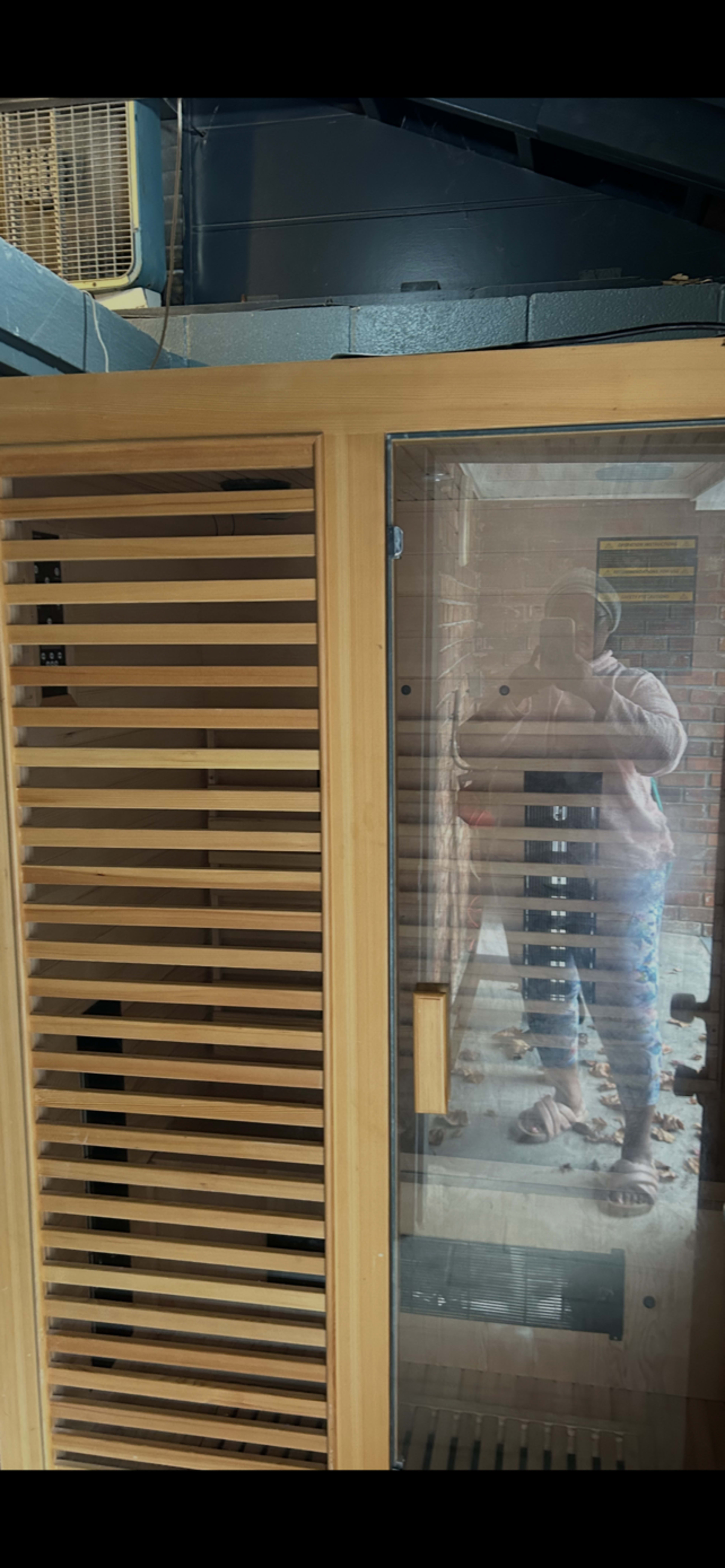 A wooden sauna with a glass door and slatted panels reflects the image of a person standing nearby.