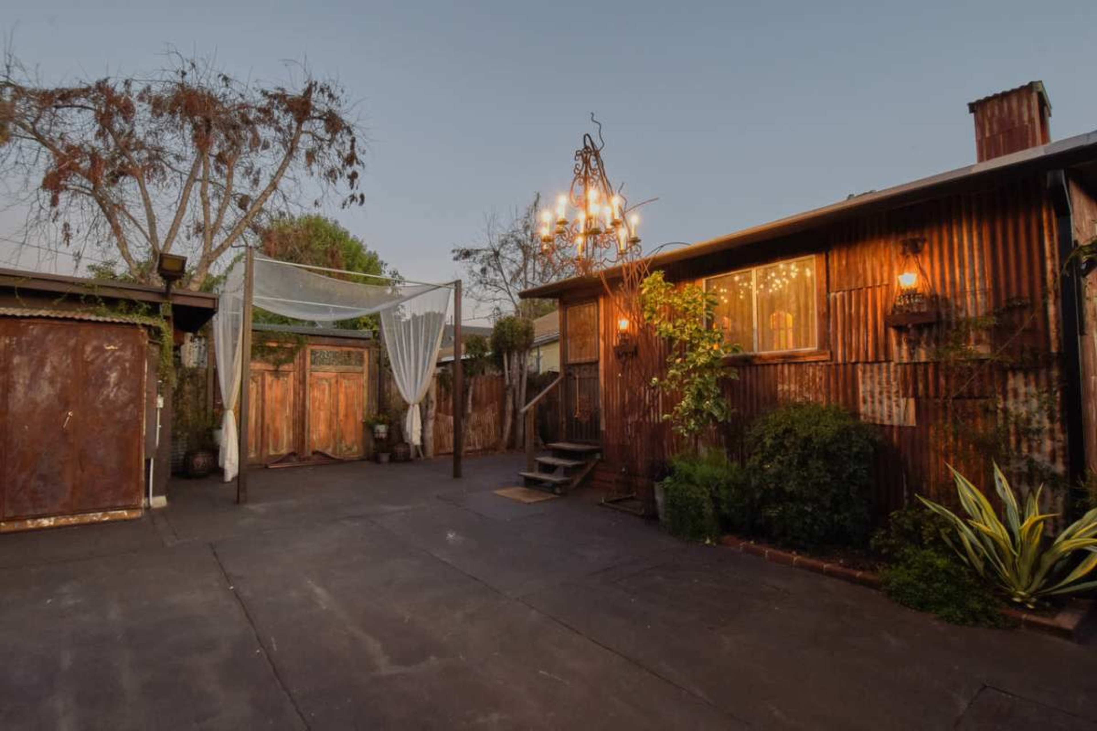 The image shows a rustic outdoor space featuring a wooden structure with a chandelier, a shaded area with drapes, and a pathway leading to a door.