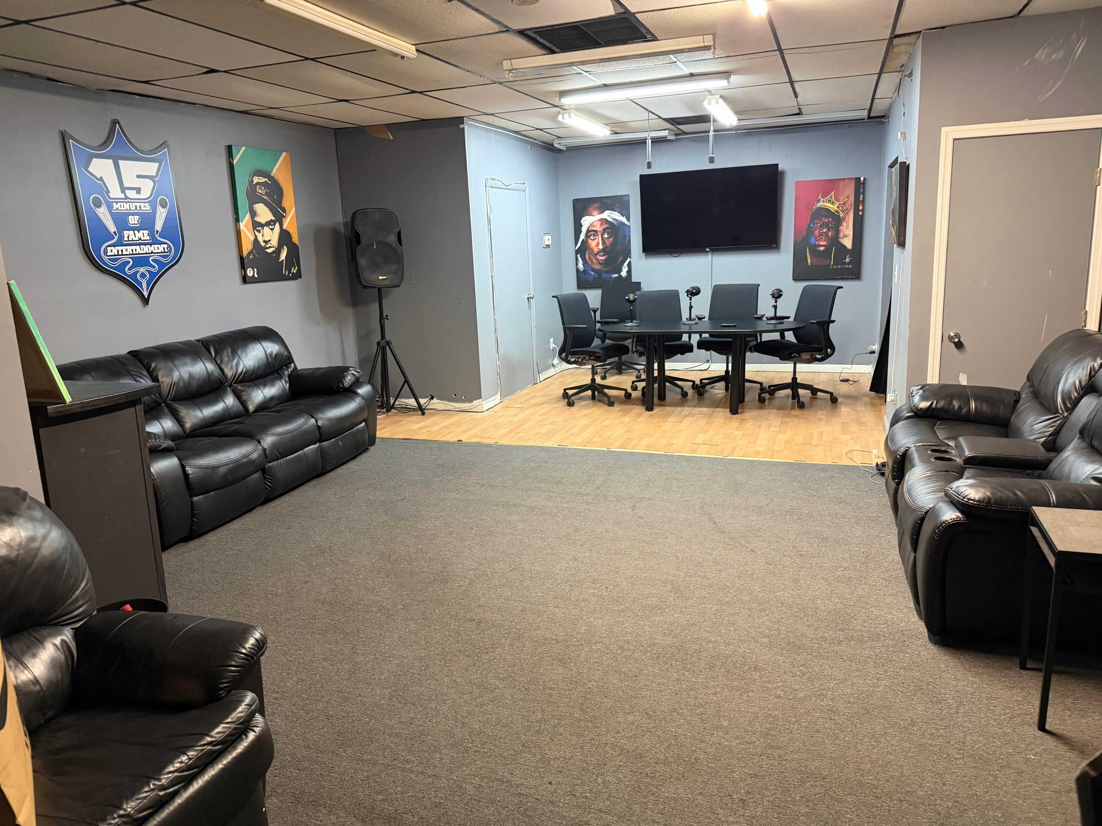 The image shows a spacious room with two black leather sofas, a central meeting table with chairs, a large screen on the wall, and framed portraits of people.