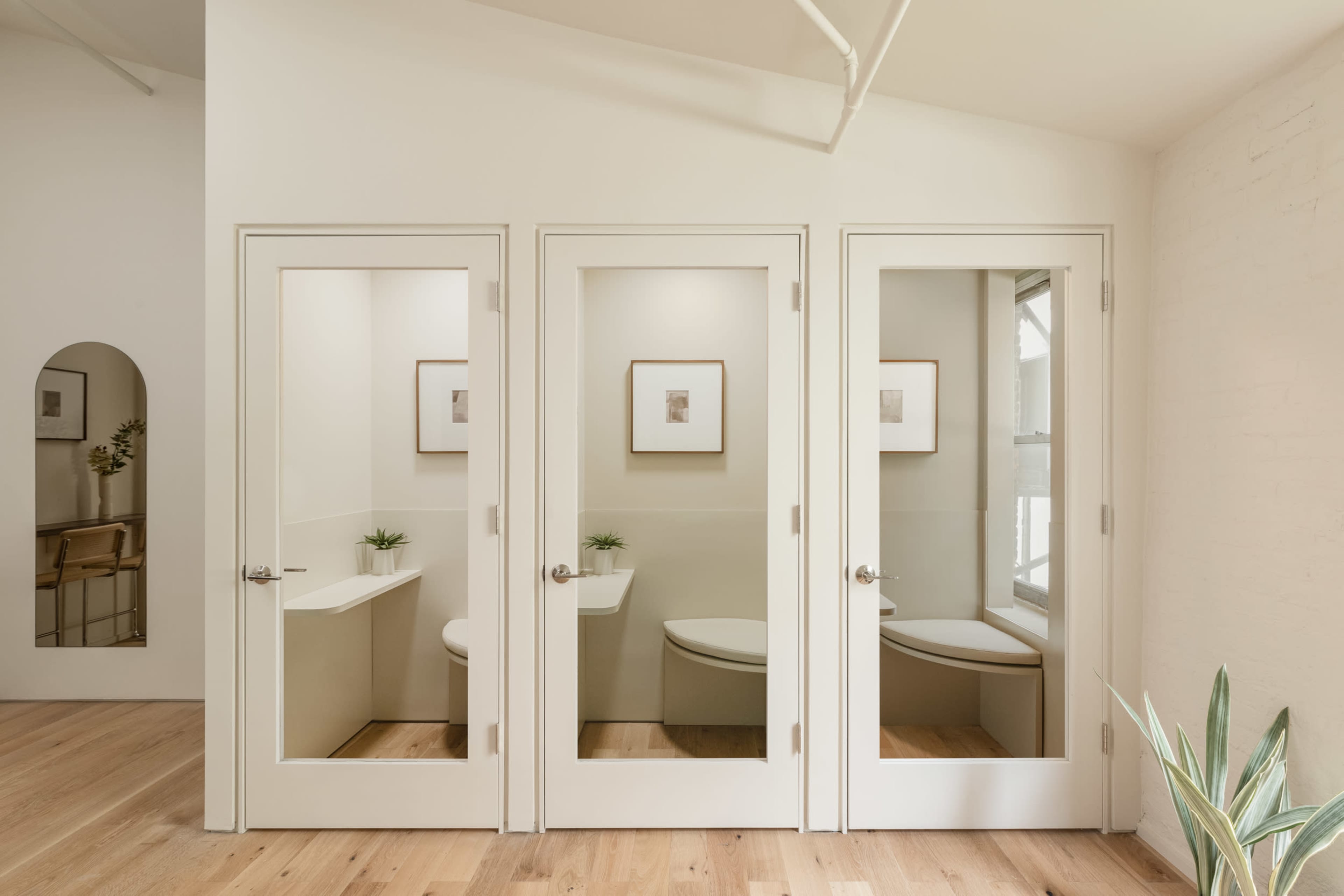 The image shows a brightly lit restroom area with three separate stalls, each featuring a clear glass door and a minimalist design.
