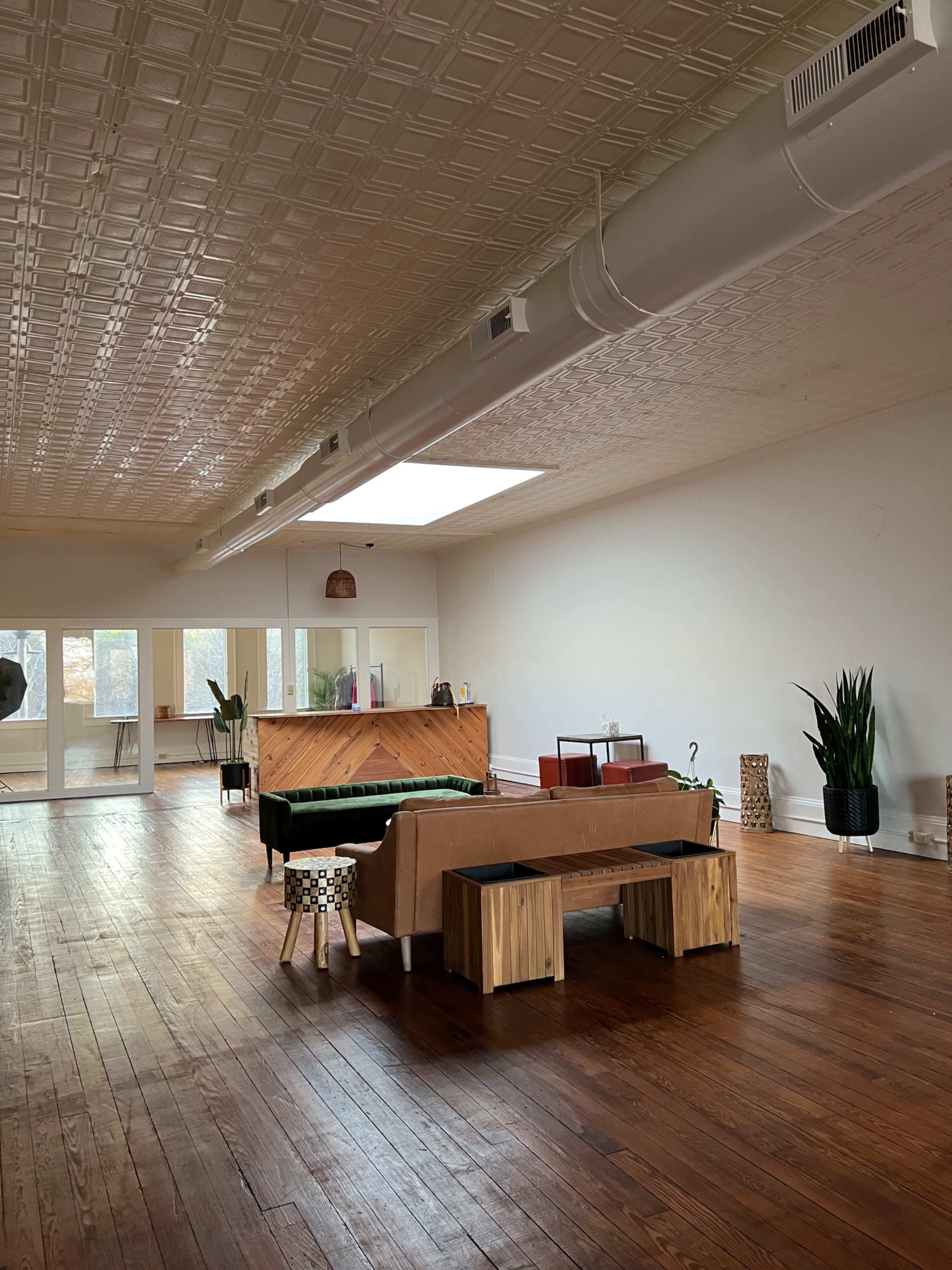 A spacious, minimalist room with wooden floors, a piano, and various plants, featuring large windows that bring in natural light.