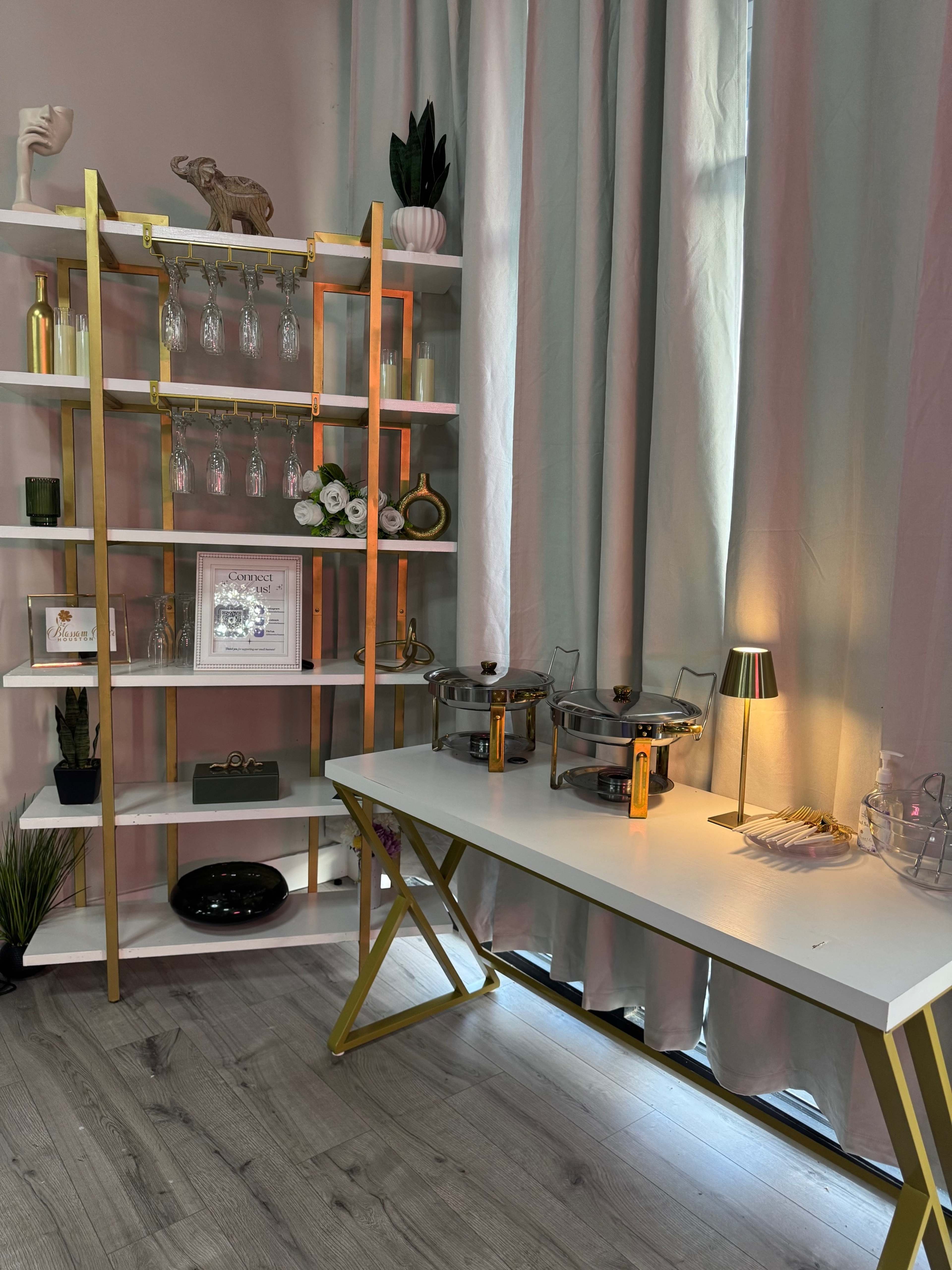 The image shows a stylish corner of a room featuring a gold-accented shelving unit with glassware, a small table with two serving dishes, and a lamp, all set against light-colored curtains.