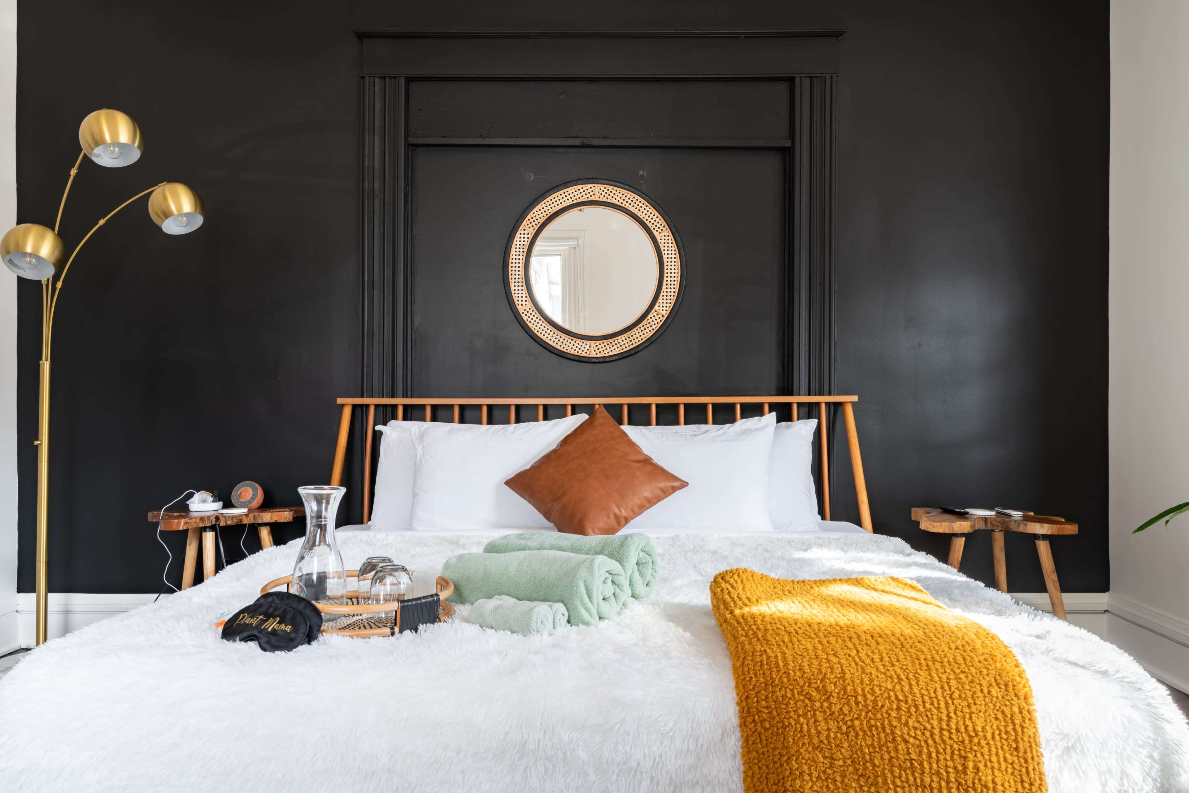 A neatly arranged bedroom features a bed with a white fur blanket, decorative pillows, and a circular mirror above a dark wall, accompanied by bedside tables and a floor lamp.