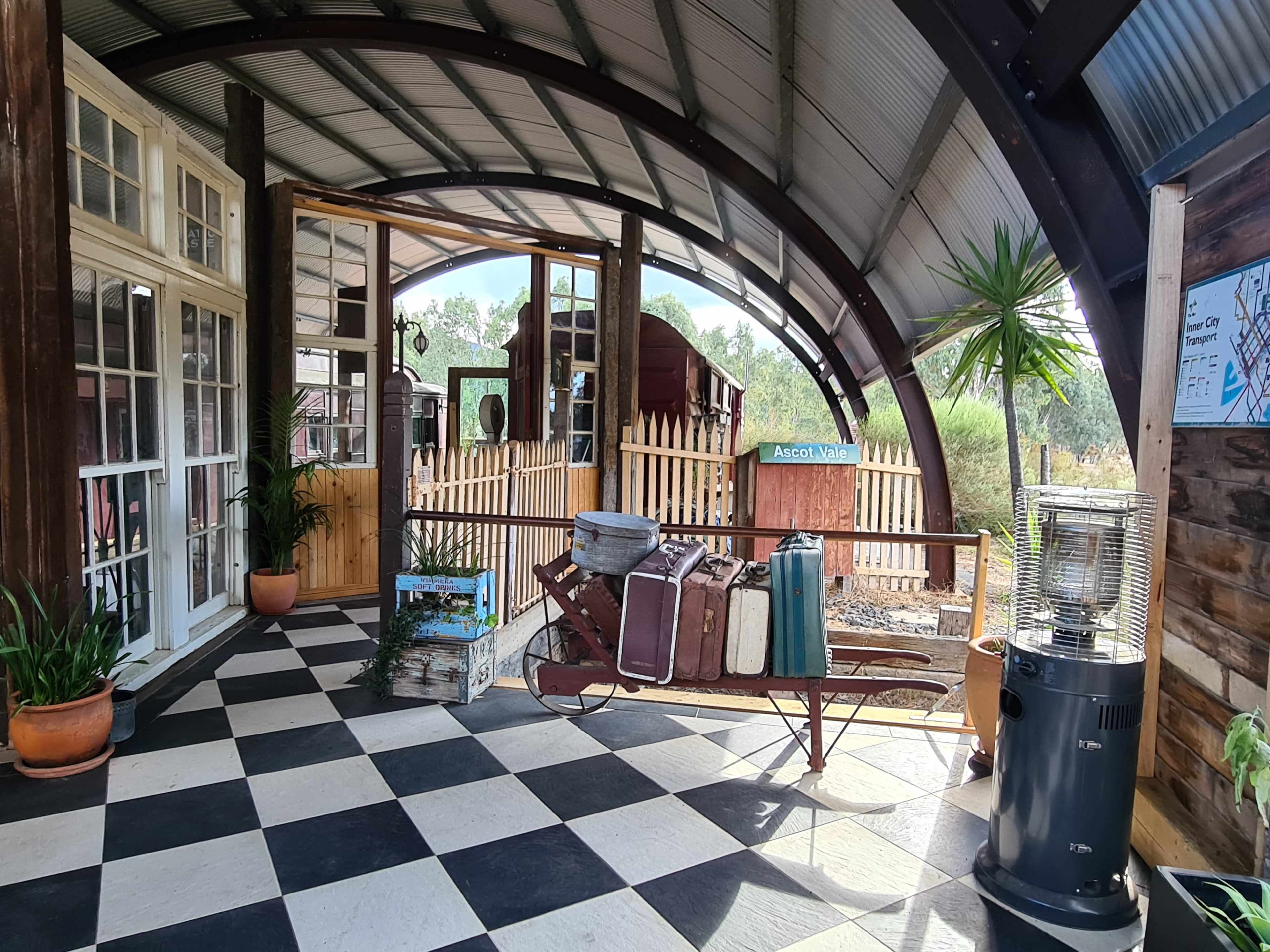The image shows an indoor area with a checkered floor, featuring a vintage sled with suitcases, large windows, and a view of a fenced outdoor space with greenery and rustic structures.