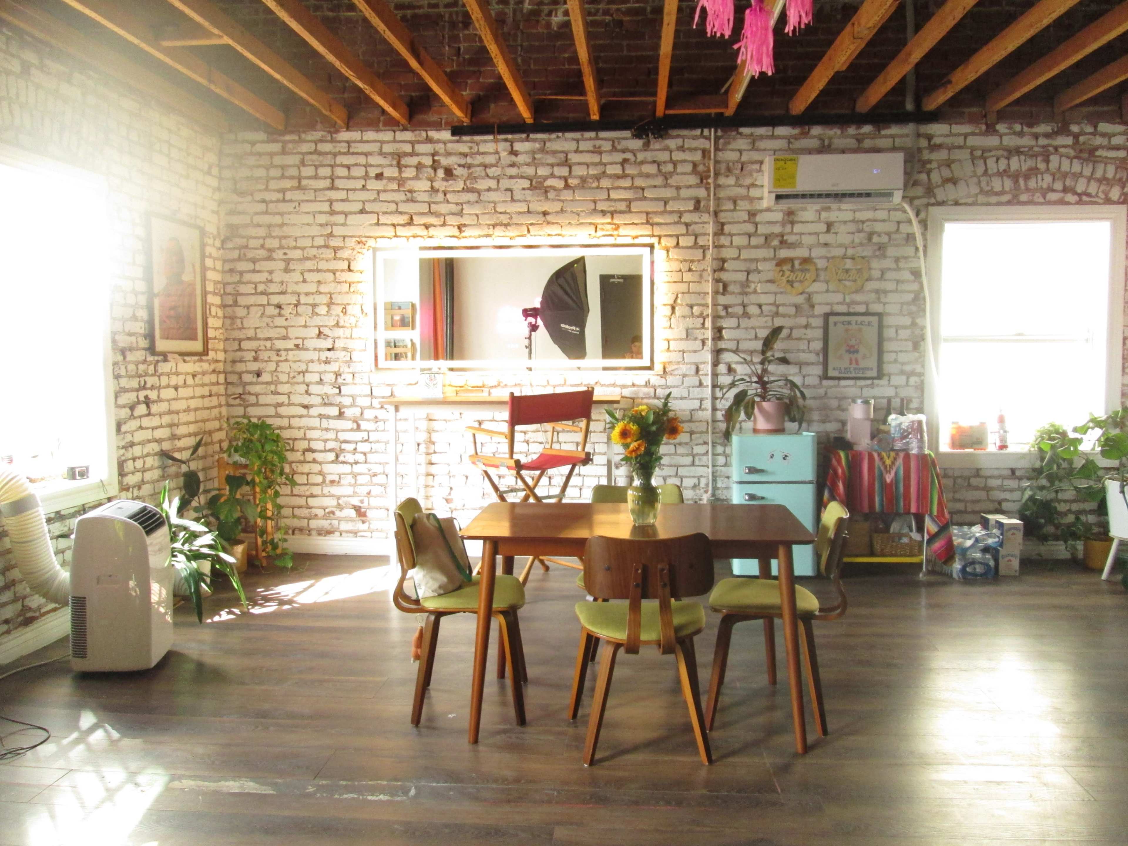 The image shows a bright, spacious interior with exposed brick walls, a wooden dining table surrounded by chairs, plants, and a mirror reflecting the room.