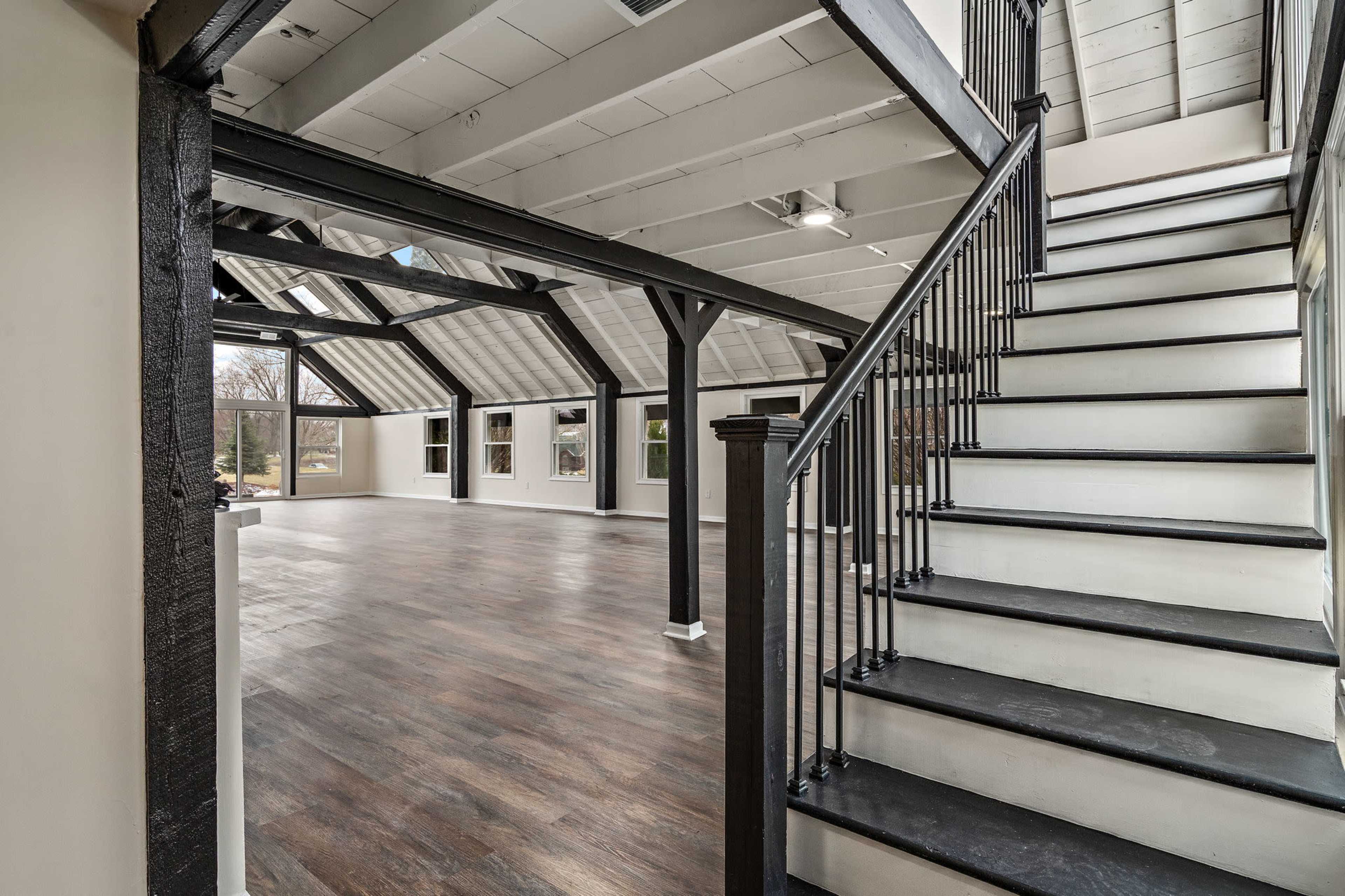 The image shows an open interior space with high ceilings, a set of black railing stairs on the right, and large windows allowing natural light to enter.