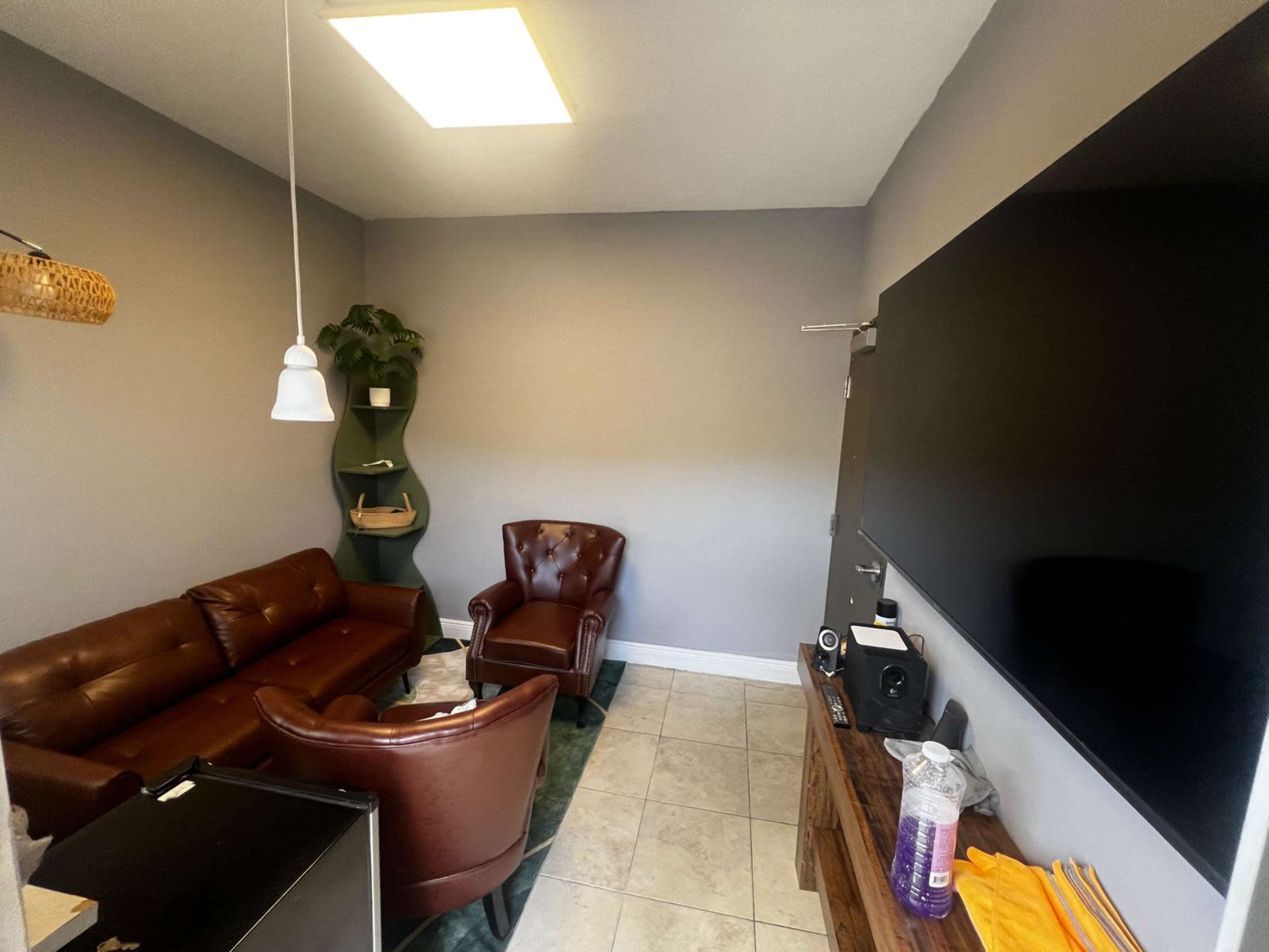 The image shows a small office space featuring a brown leather sofa, two armchairs, a television on the wall, and a potted plant in the corner.