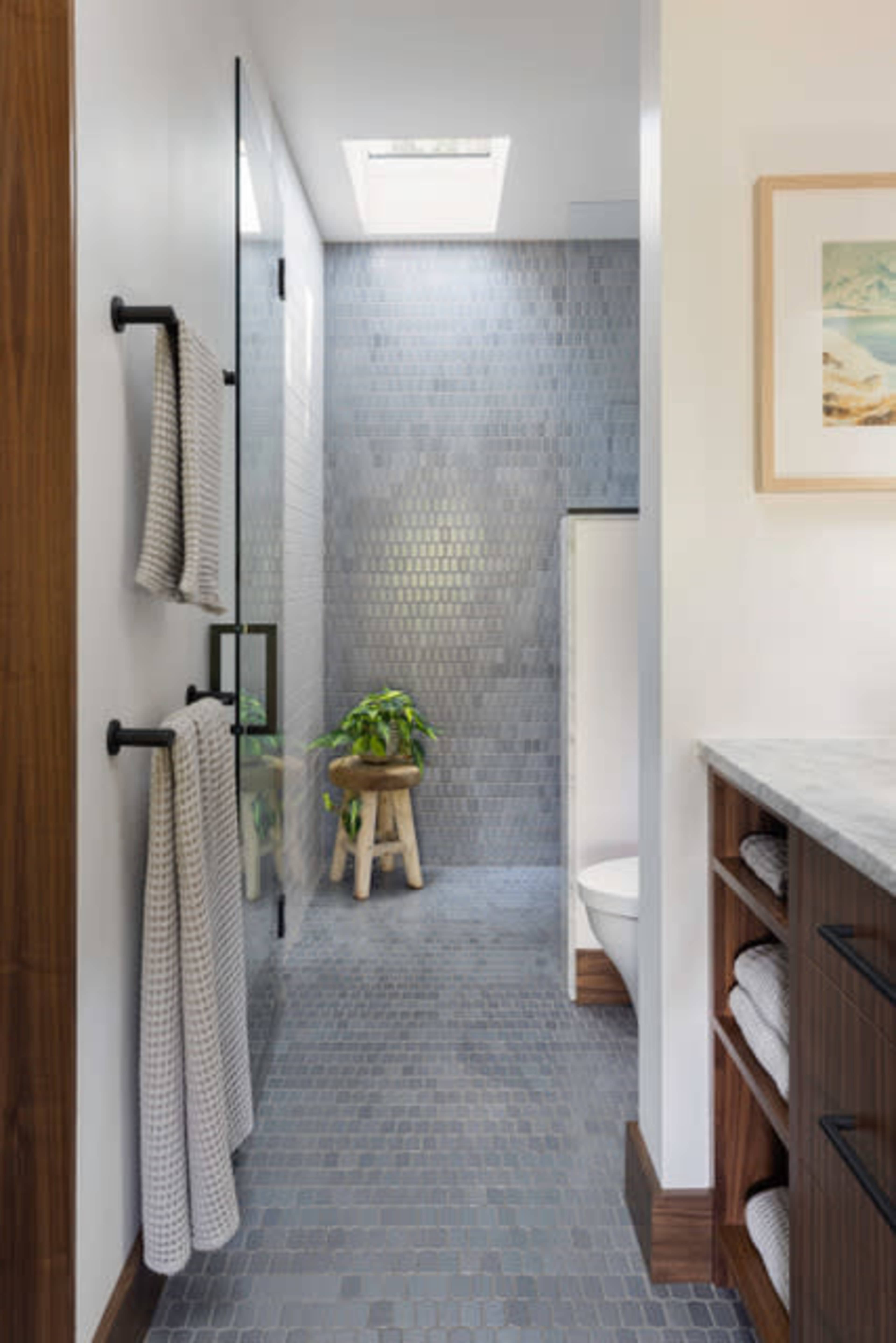 A modern bathroom features a glass shower enclosure, gray tiled walls, a skylight, and a wooden stool with a potted plant.