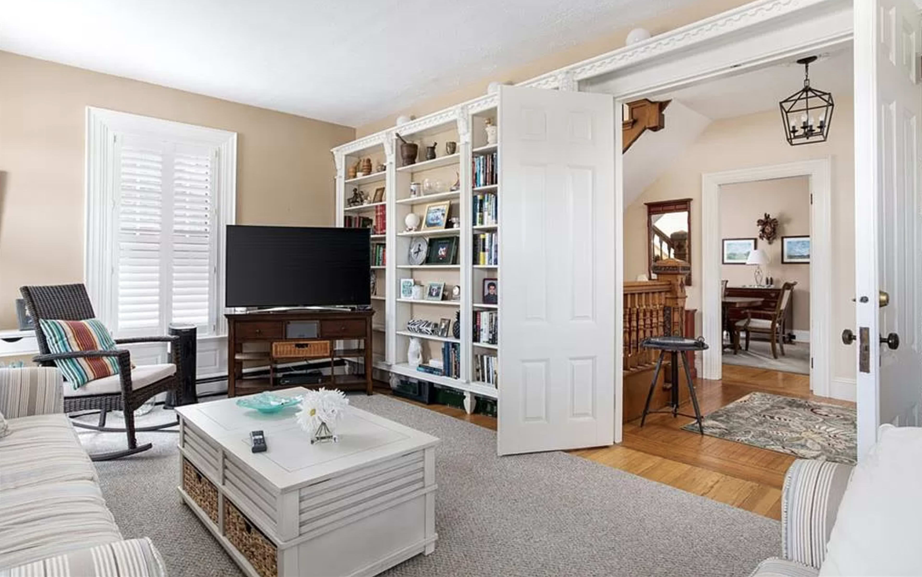 The image shows a cozy living room featuring a bookshelf, a television, and a coffee table, connected to a dining area through an open doorway.