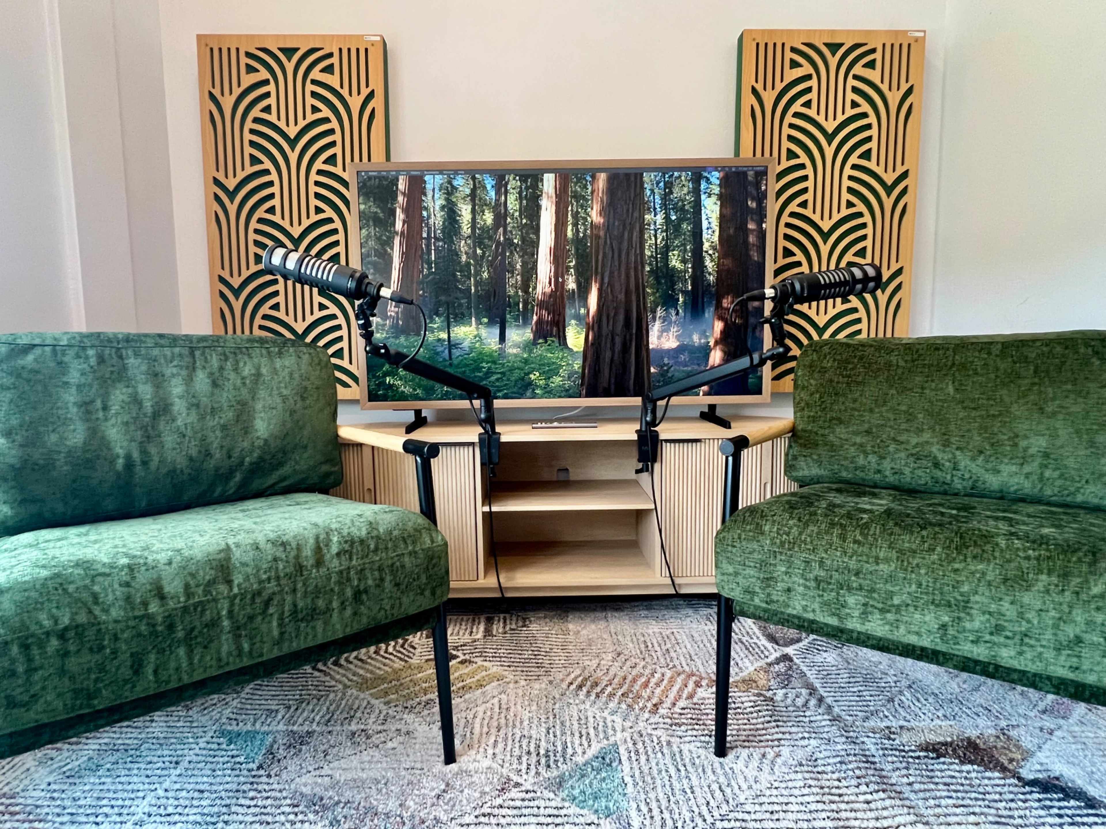 The image shows a cozy living room setup featuring two green velvet chairs positioned in front of a television displaying a forest scene, with two microphones mounted on stands nearby.
