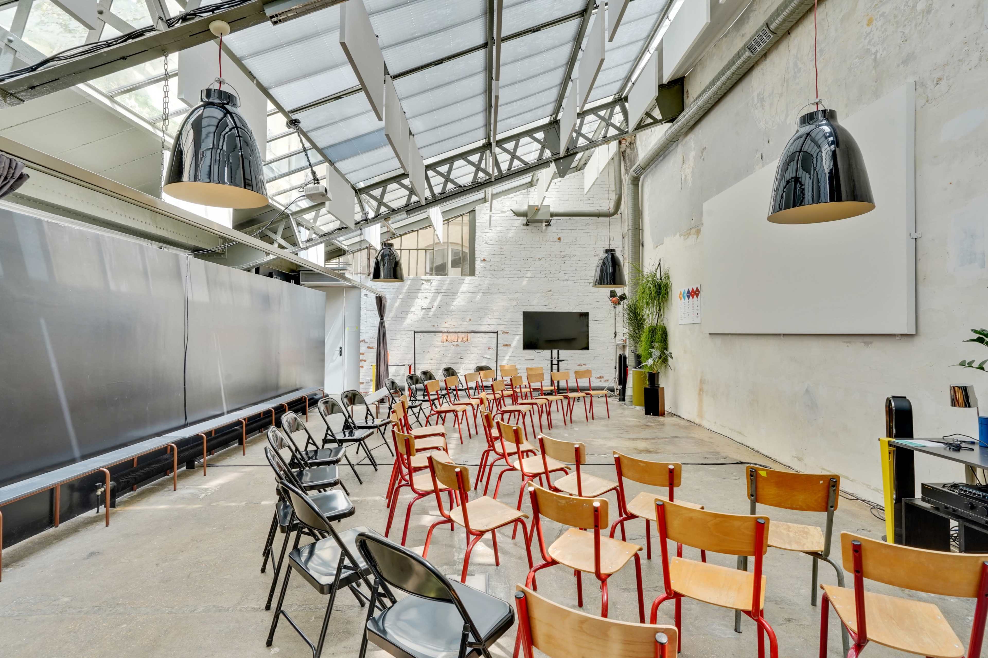 A spacious room with rows of colorful chairs arranged for a meeting, featuring large windows and industrial design elements.