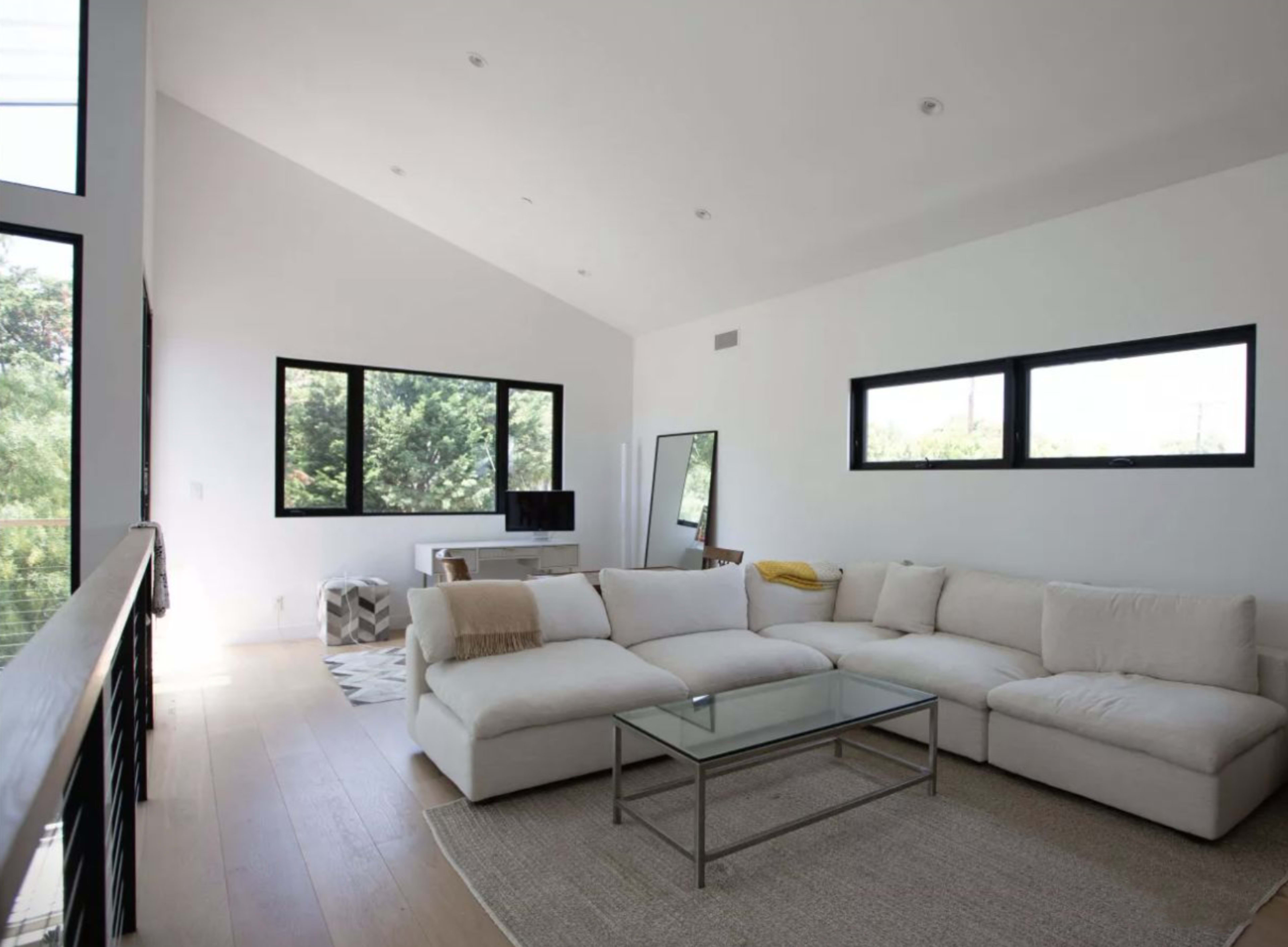 A spacious living room with a large white sectional sofa, a glass coffee table, and modern windows revealing greenery outside.