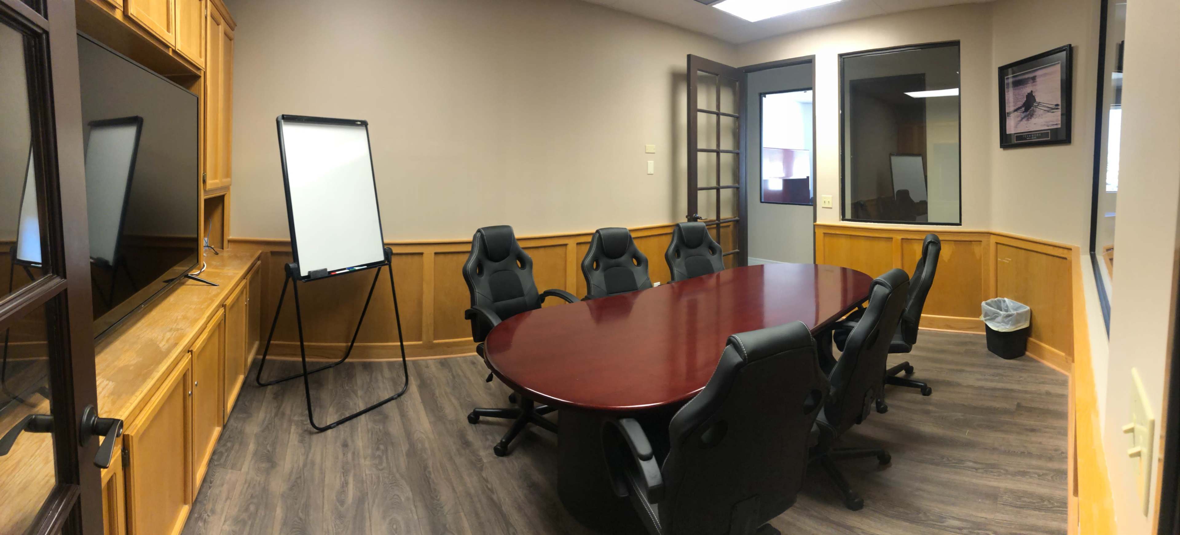 A conference room features a large oval table surrounded by six black chairs, a whiteboard on an easel, a television on the wall, and wooden paneling.