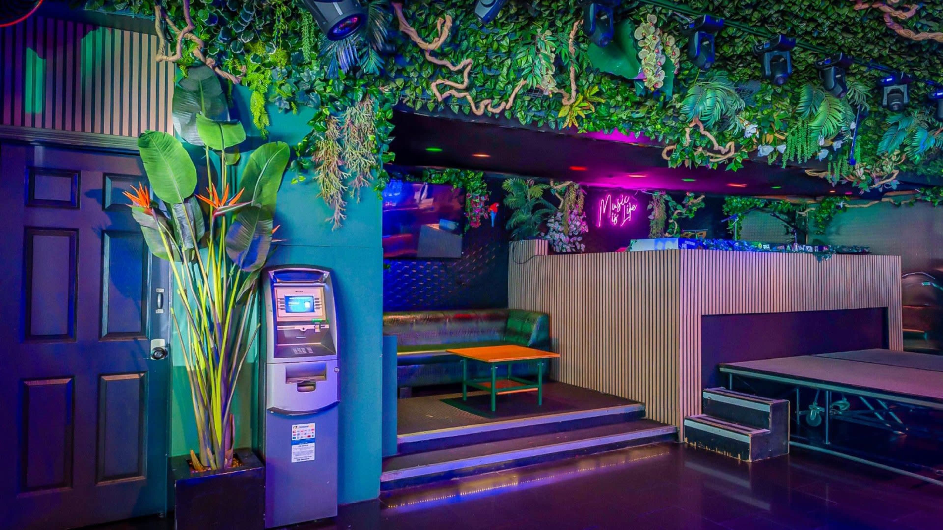 A vibrant lounge space with green foliage decor, a seating area, a small table, and an ATM machine near the entrance.