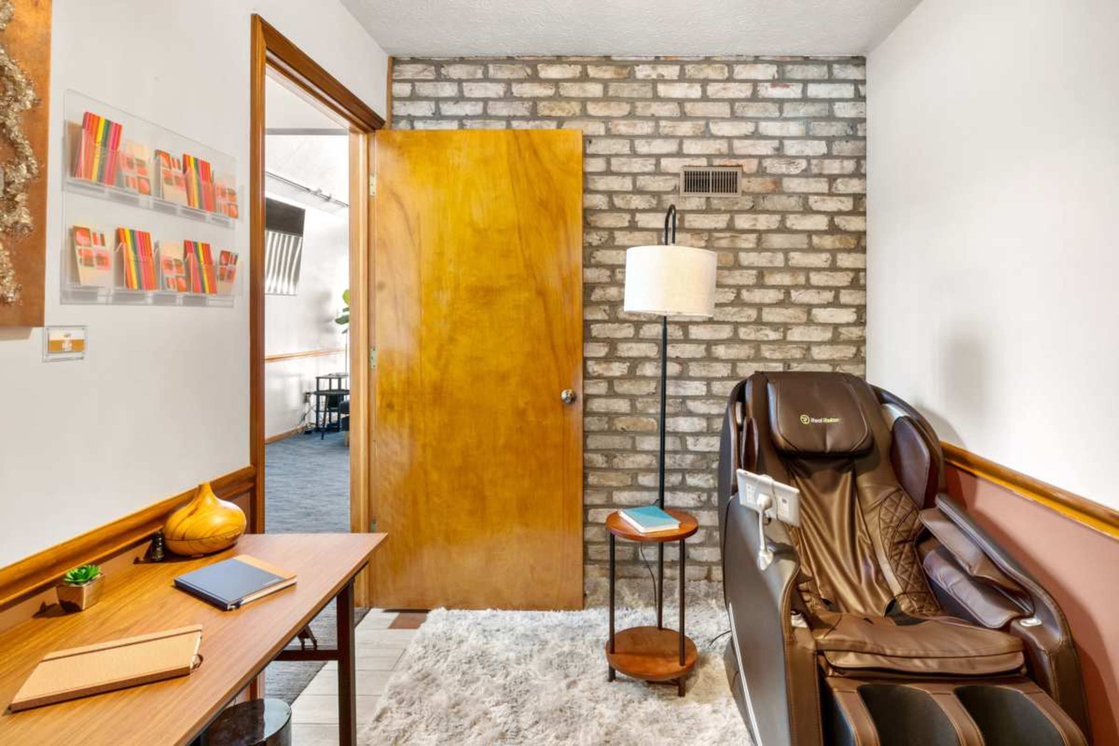 The image shows a cozy room featuring a massage chair, a wooden door, a small round table with a lamp, and a decorative wall with a brick texture.