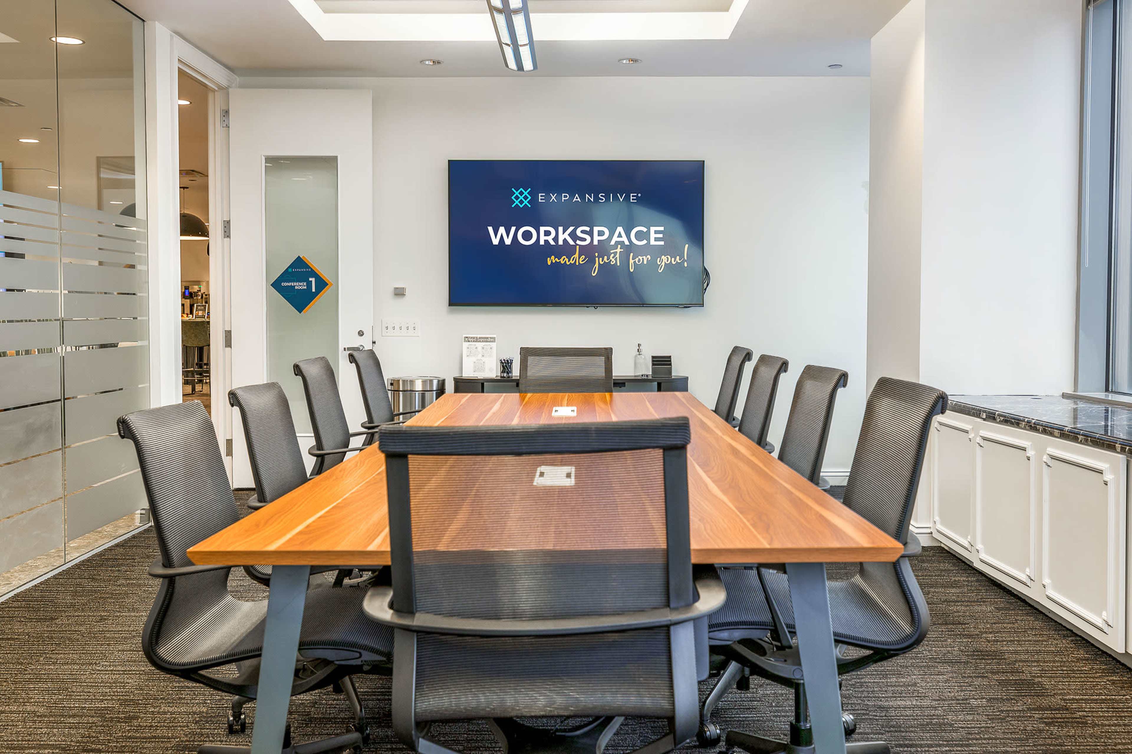 A large conference room features a wooden table surrounded by ergonomic black chairs, with a display screen mounted on the wall that reads "WORKSPACE made just for you!"