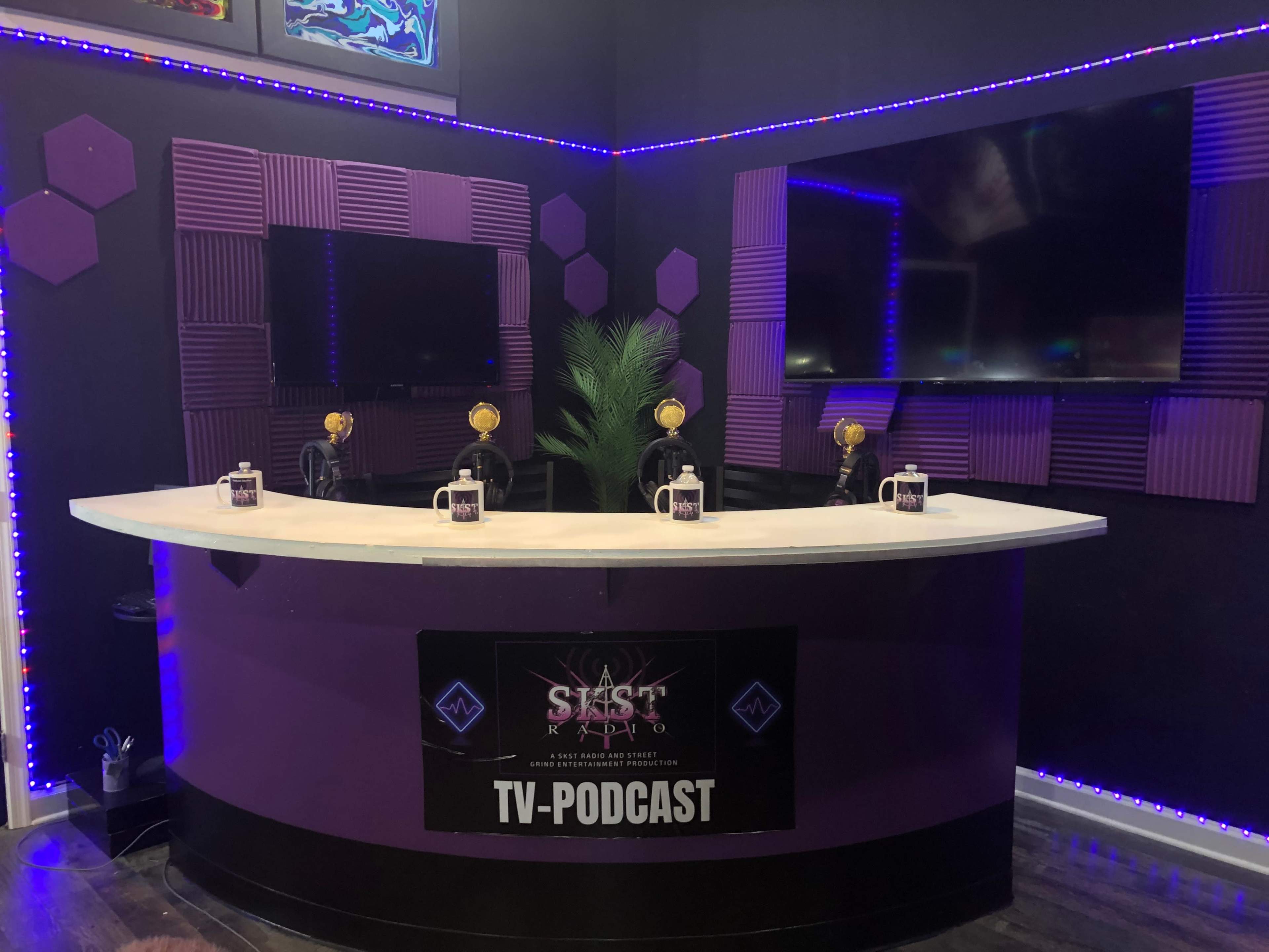 The image shows a modern podcast studio with a circular white desk, four microphones, and two large screens on a purple and black wall.