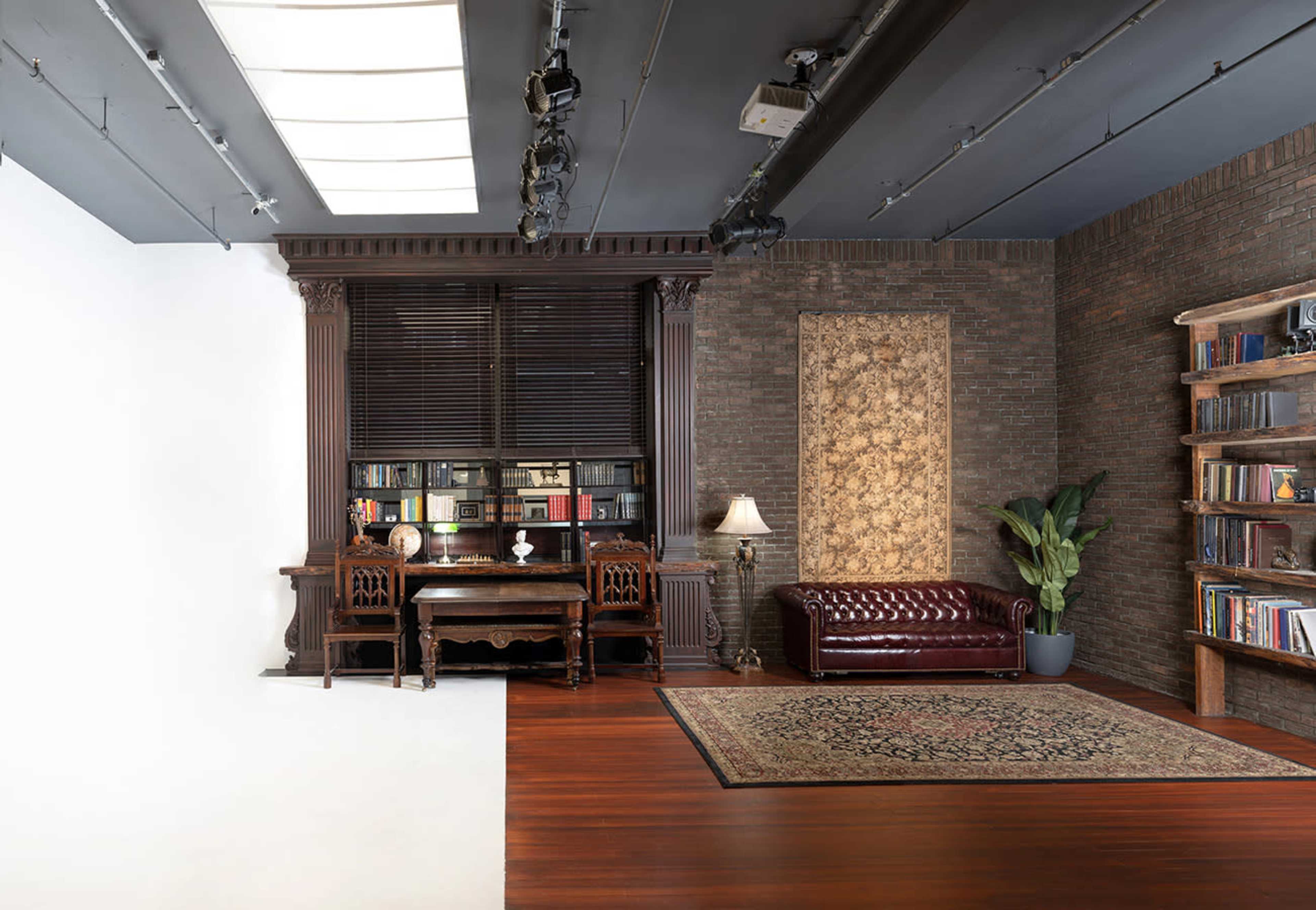 A spacious interior with a dark wooden desk, a leather sofa, a bookshelf filled with books, and a potted plant, set against a backdrop of a textured brick wall.