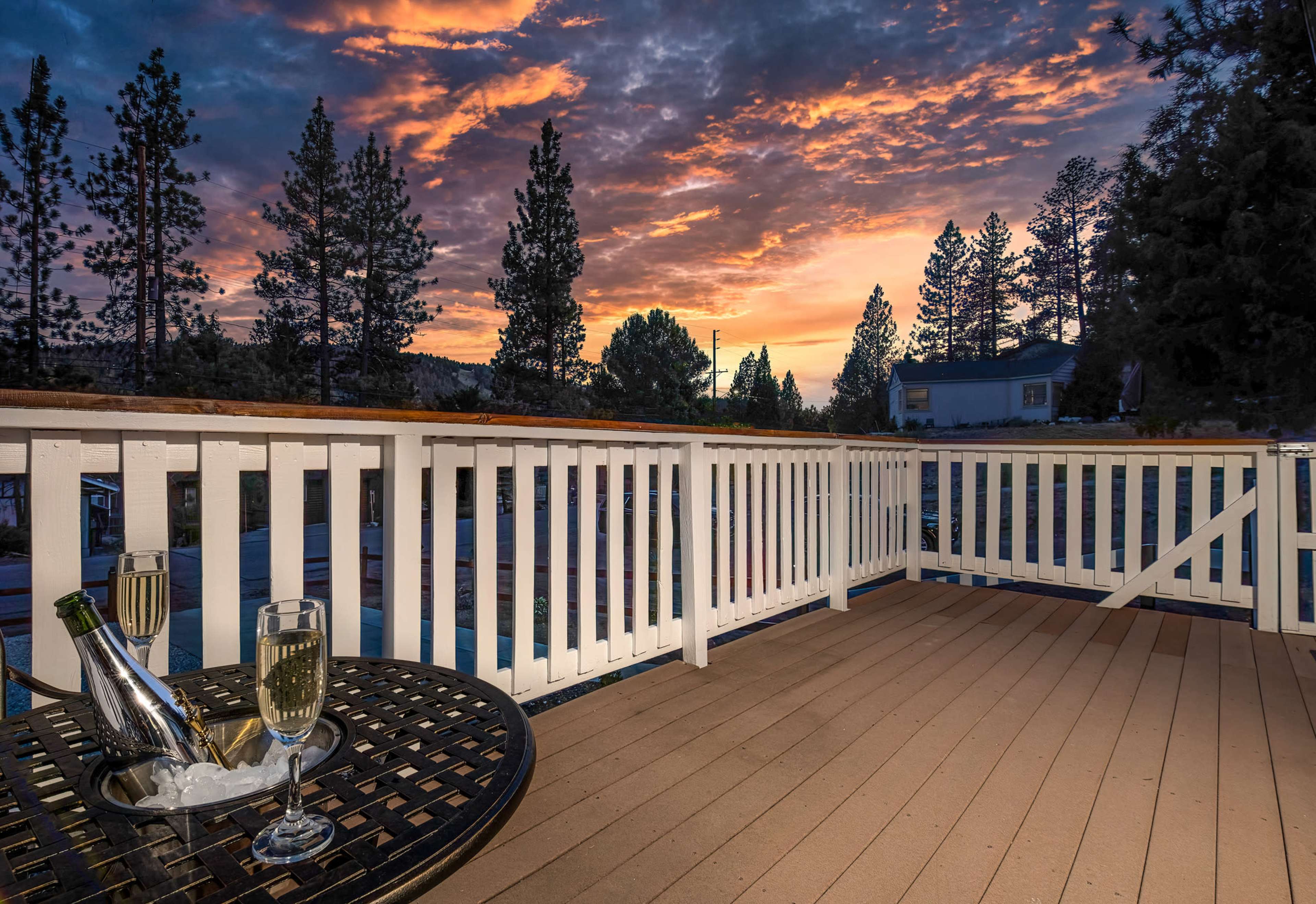 A bottle of champagne and two glasses sit on a black table on a deck, overlooking a vibrant sunset with trees in the background.