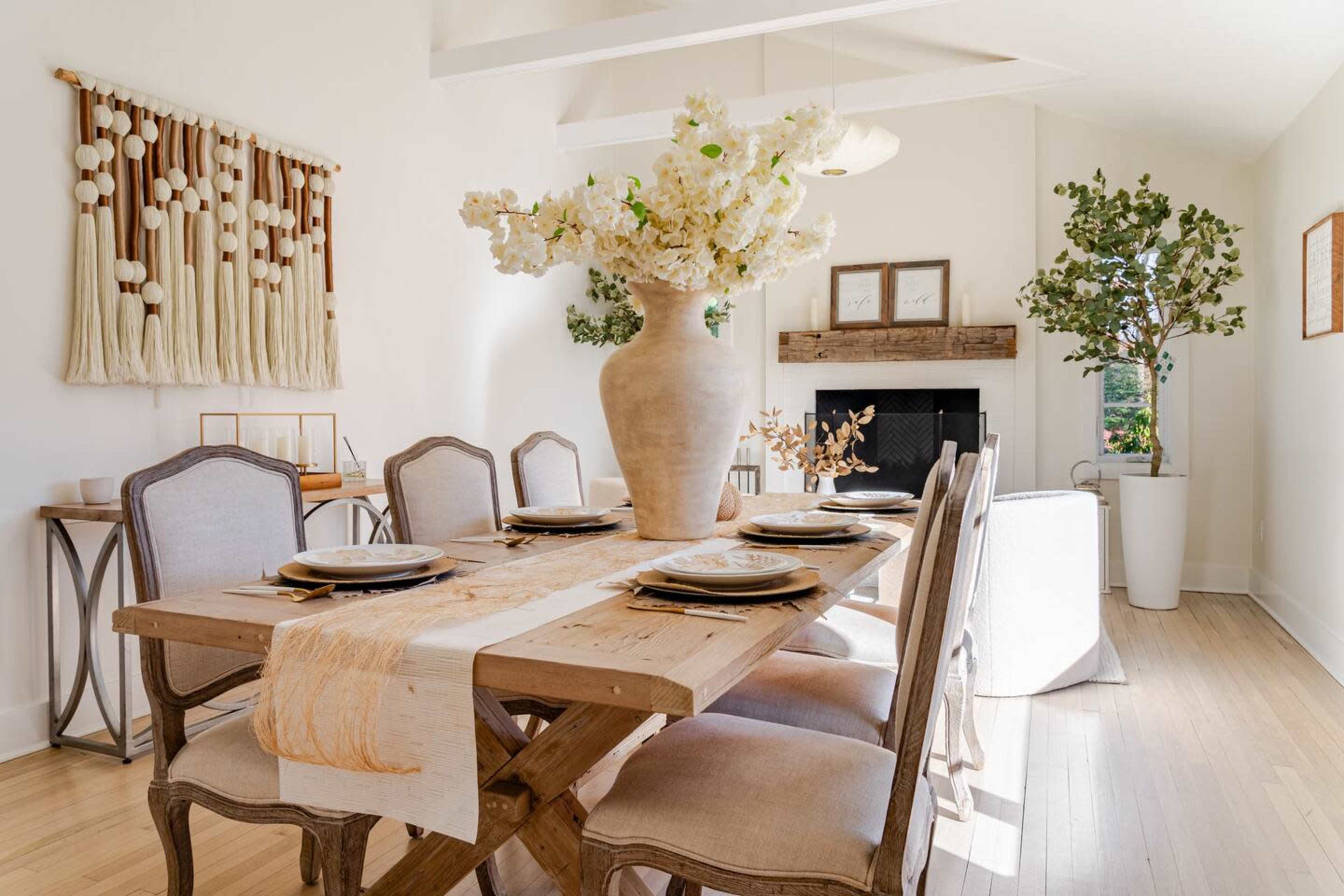 A spacious dining room features a wooden table set with plates and a large vase of artificial flowers, surrounded by chairs and decorative plants.