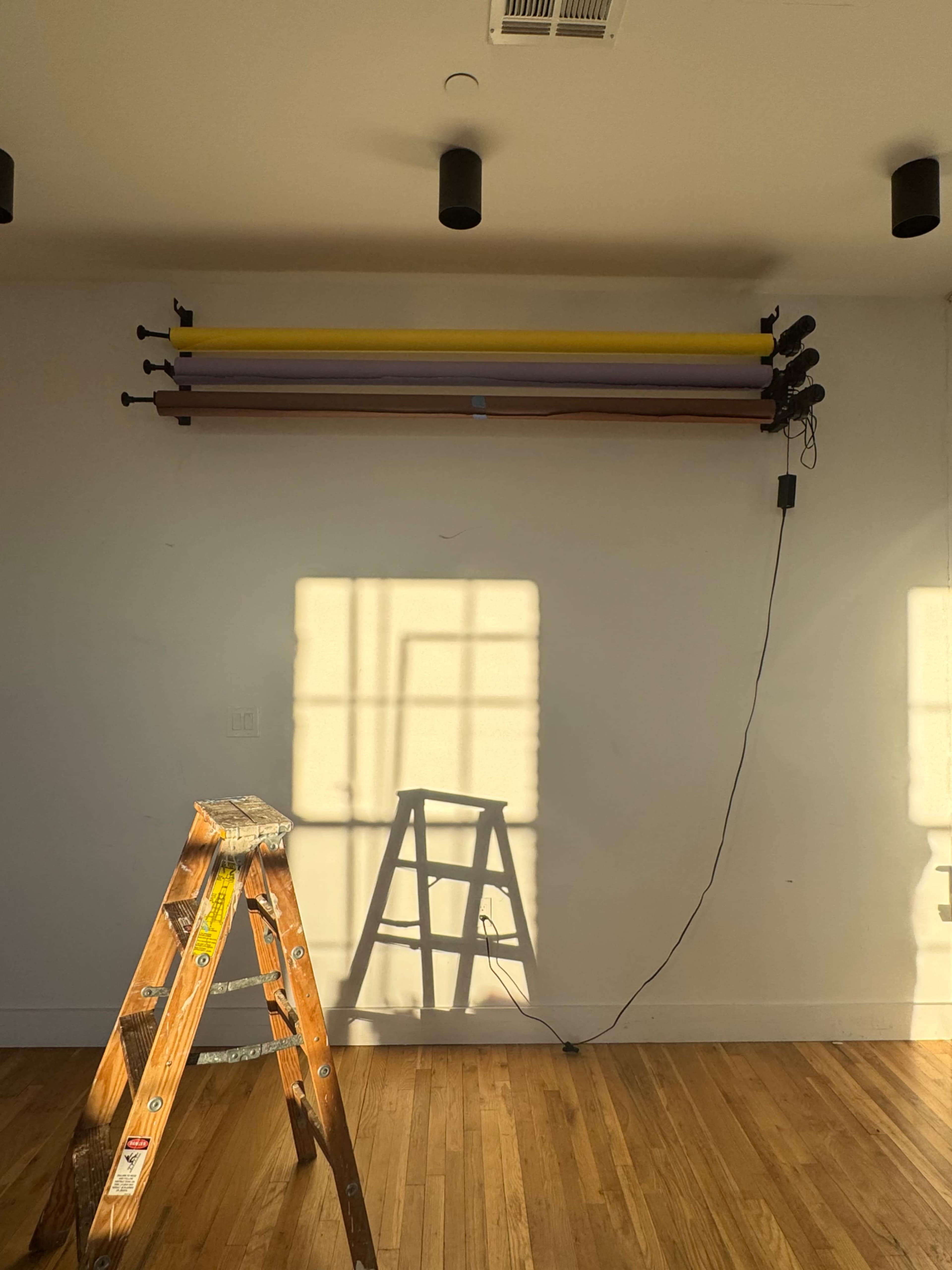 A wooden ladder stands in front of a wall with four colored backdrop poles mounted above, casting a shadow on the floor from sunlight filtering through a window.