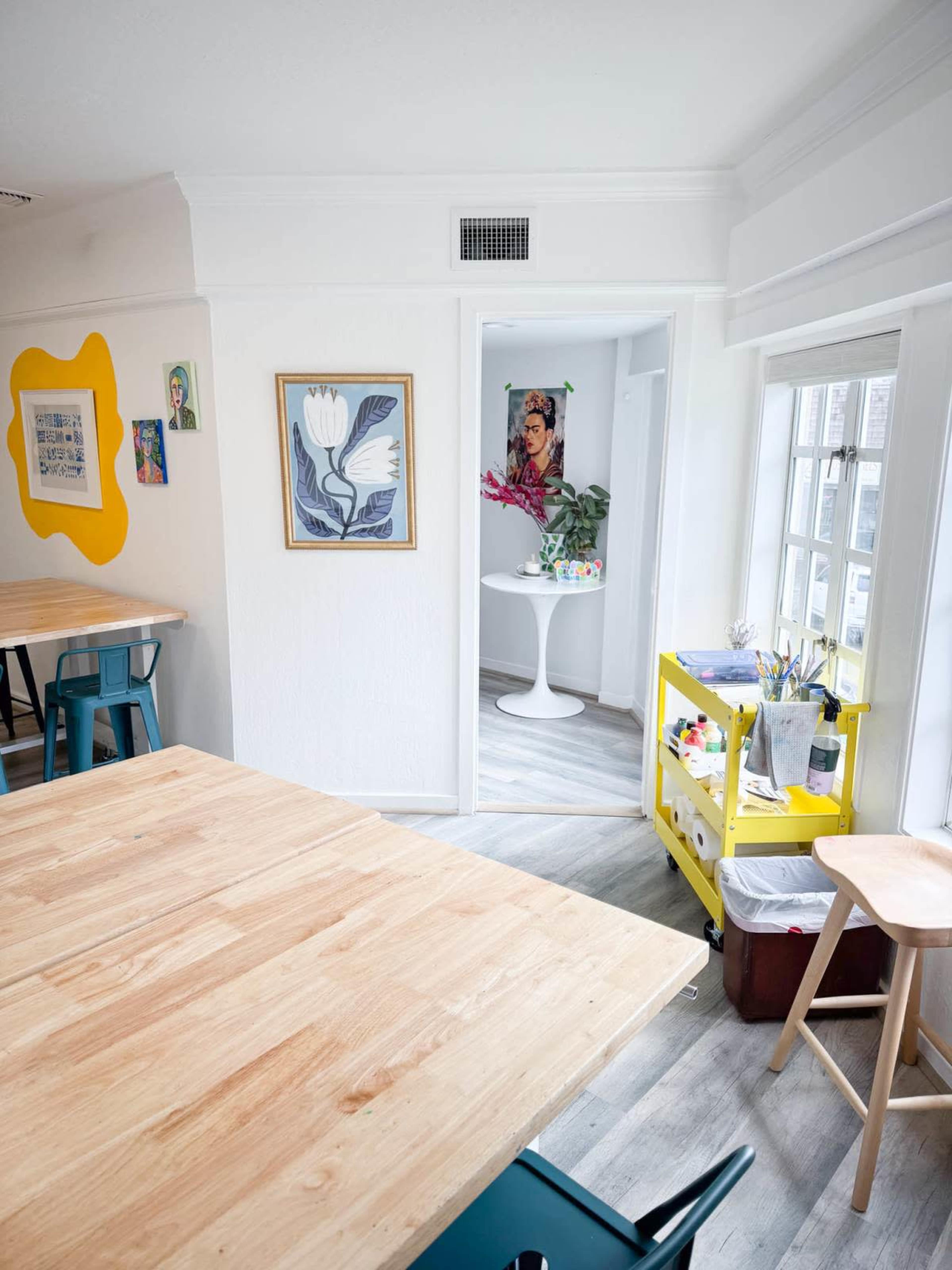 A bright and airy room features wooden tables, colorful artwork on the walls, and a small cart with art supplies near a doorway leading to another space.