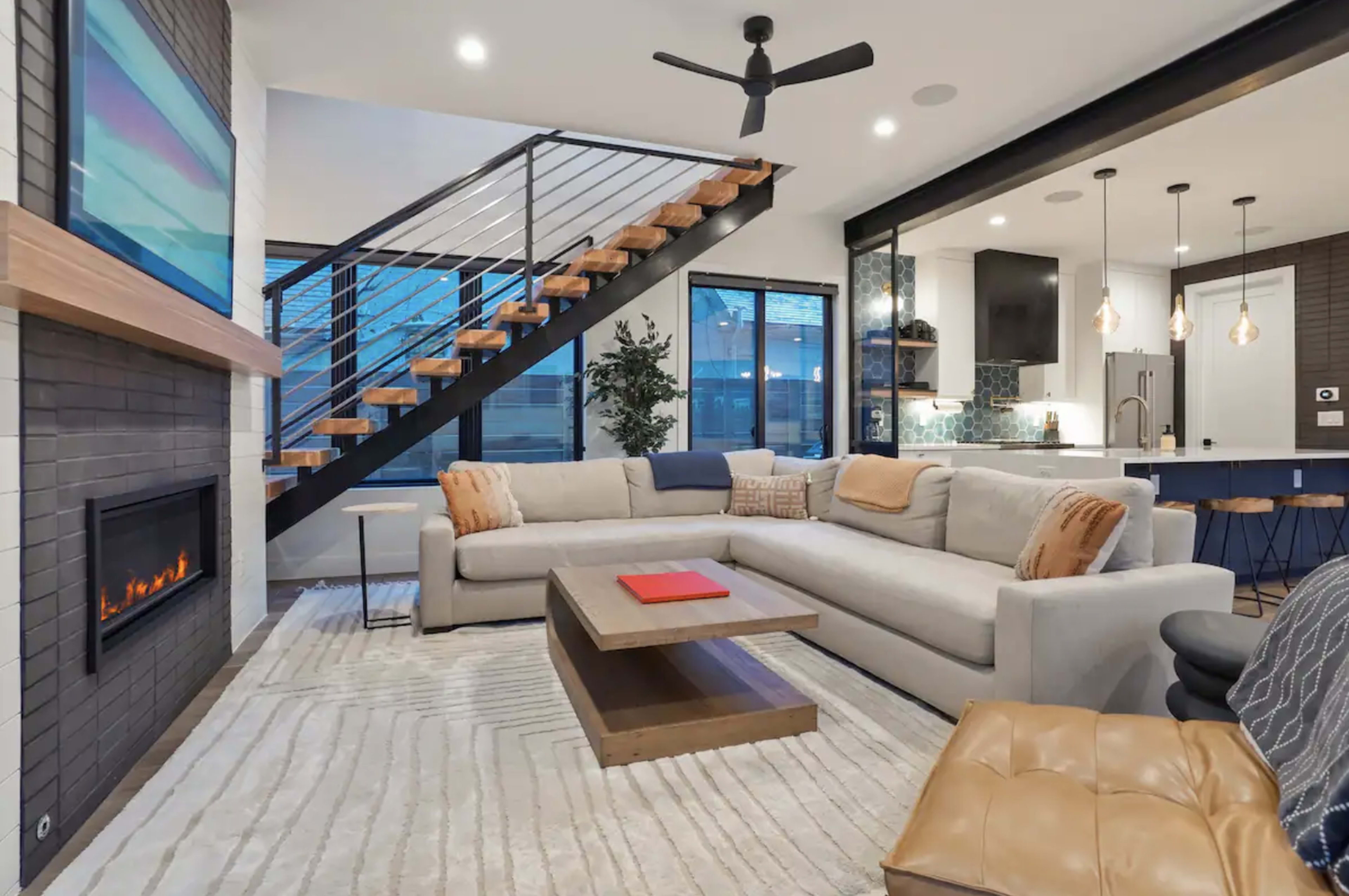 A modern living room with a large sectional sofa, a coffee table, a fireplace, and an open staircase leading to an upper level.