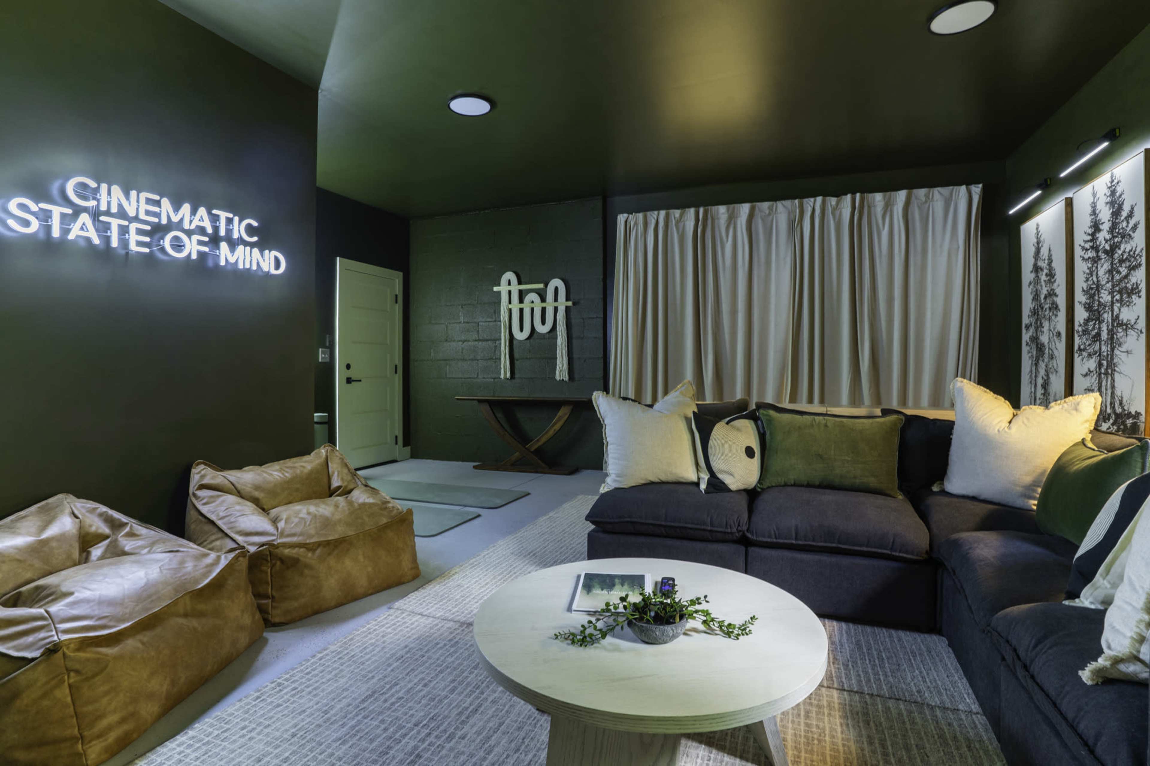 The image depicts a cozy living room with dark green walls, a large sectional sofa, bean bag chairs, and a decorative table, illuminated by wall-mounted signs and soft lighting.