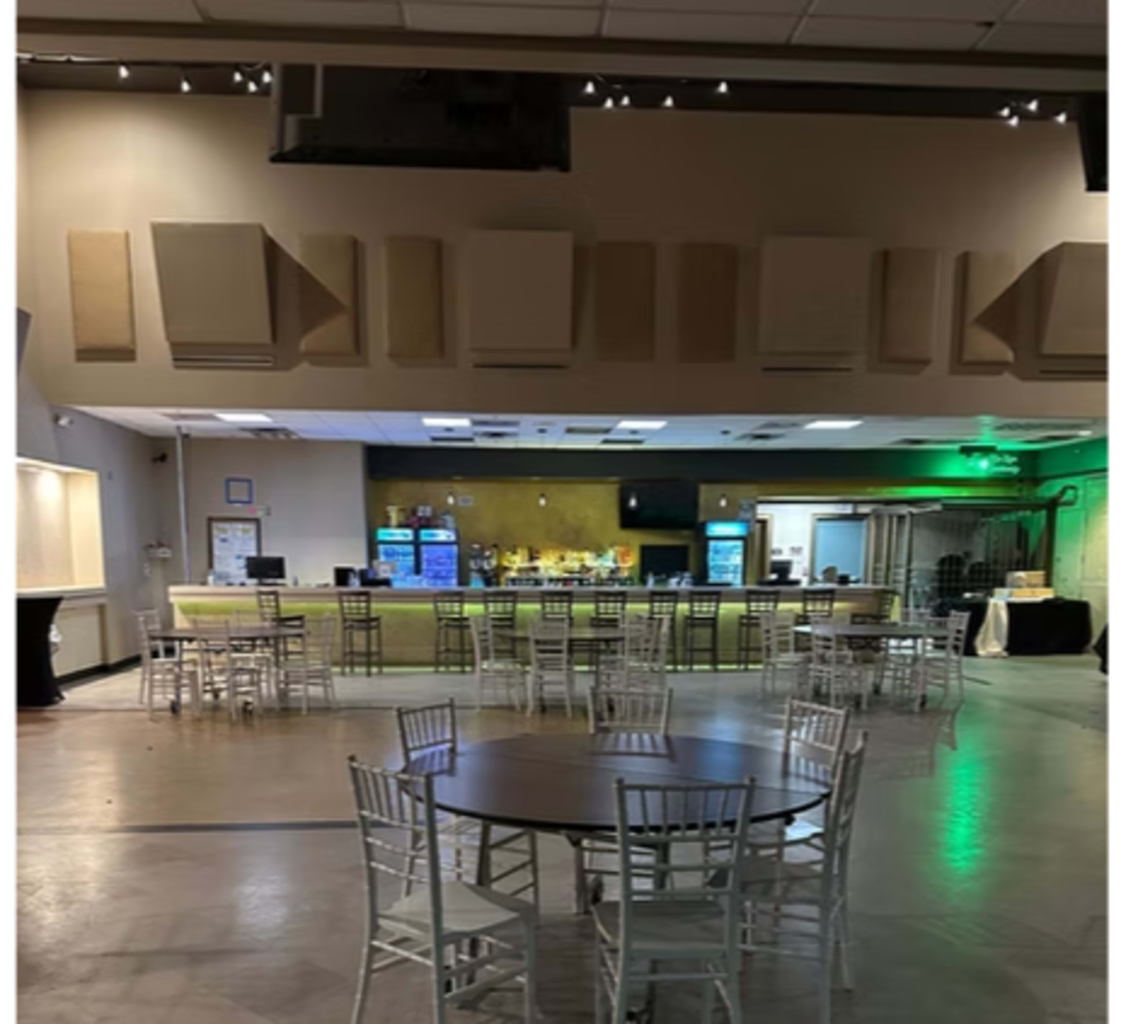 The image shows a spacious interior of a venue with a bar in the background, and several tables and chairs set up in the foreground.