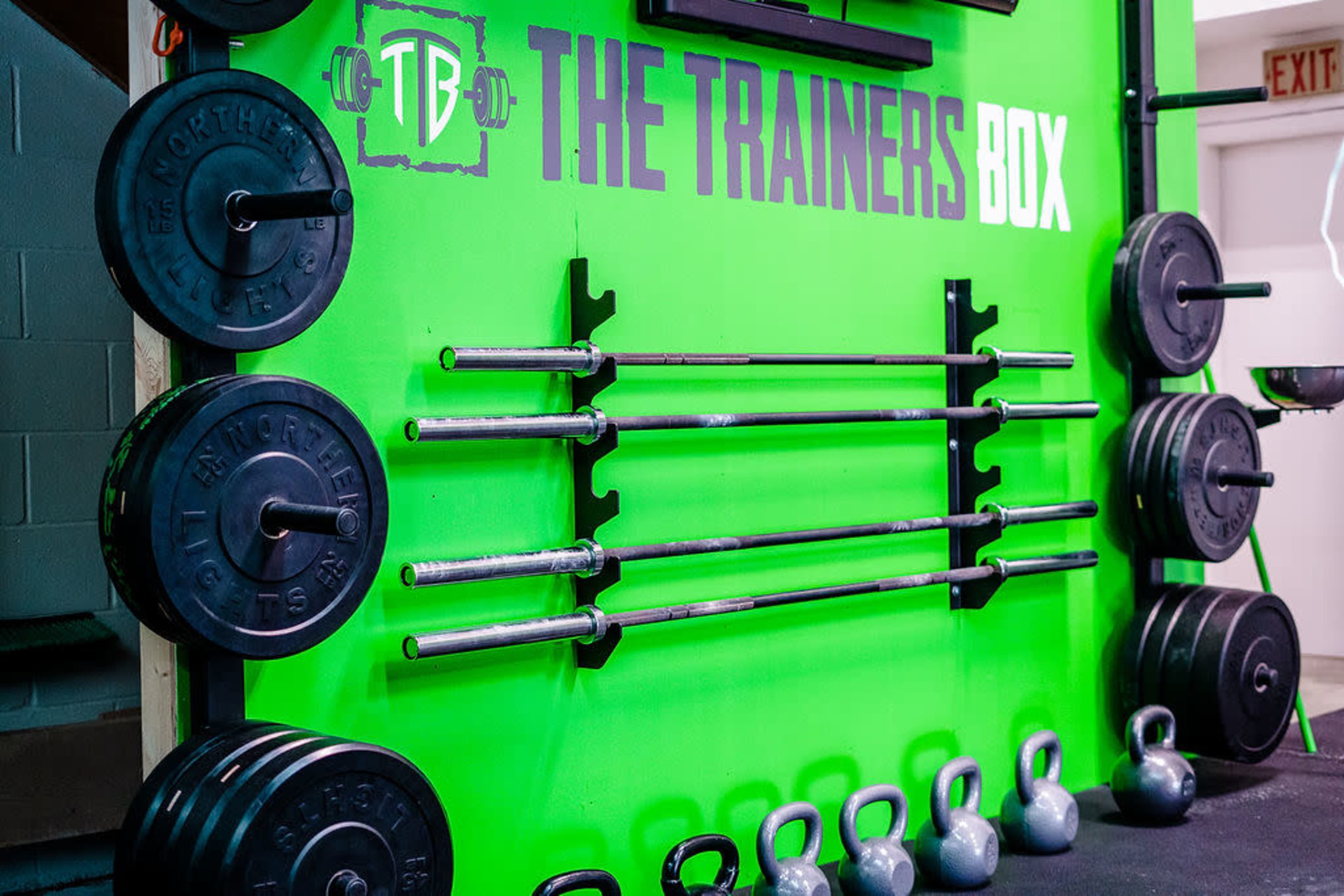 A fitness studio wall with various weight plates, barbells, and kettlebells arranged neatly against a bright green background featuring the text "THE TRAINERS BOX."