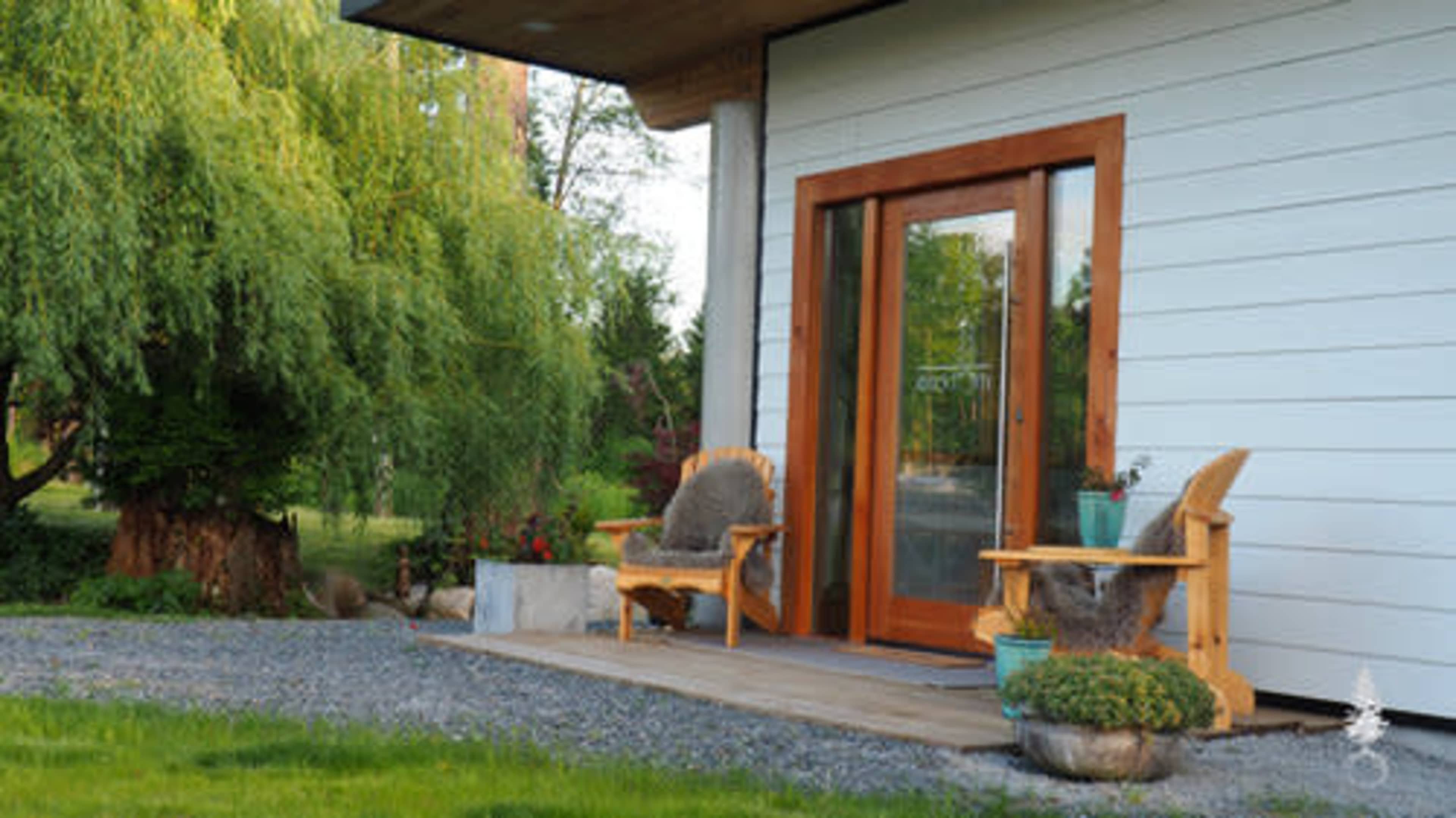 A wooden porch with two chairs and a potted plant sits beside a grassy area and trees.