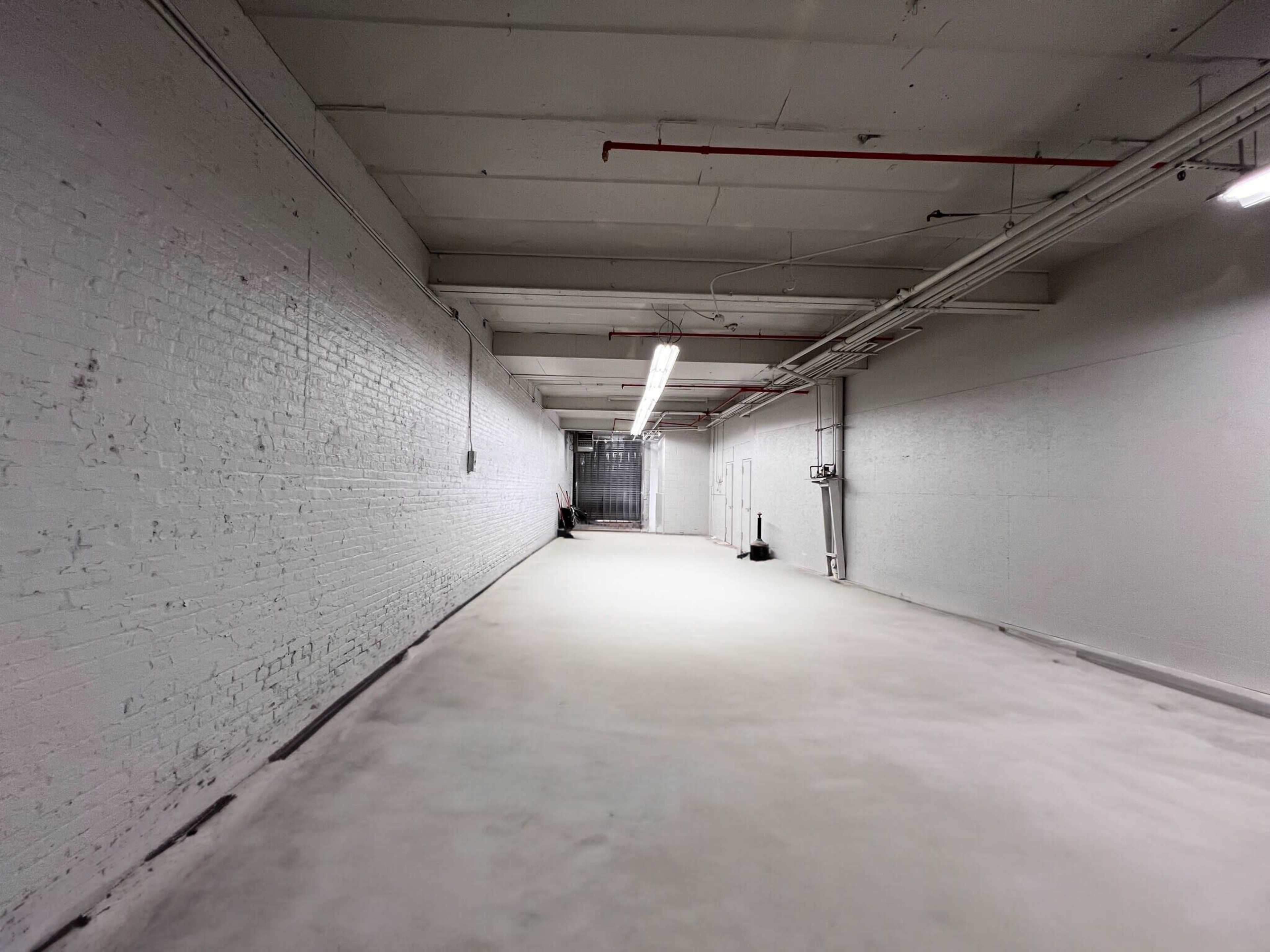 The image shows a long, empty corridor with brick walls and a concrete floor, illuminated by overhead lights.